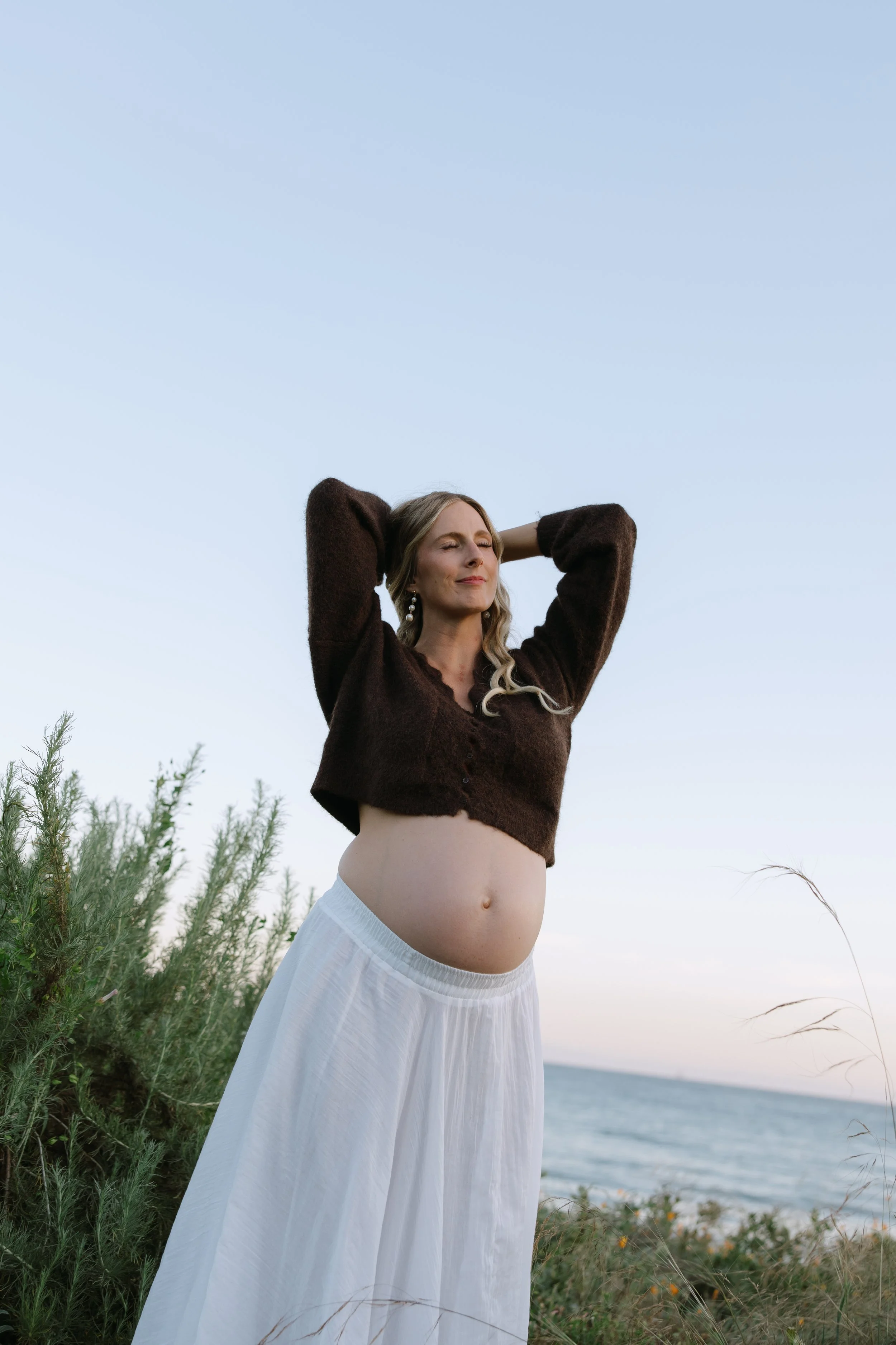 A pregnant woman standing outdoors near the ocean, with her hands behind her head, eyes closed, wearing a dark sweater and a flowing white skirt.