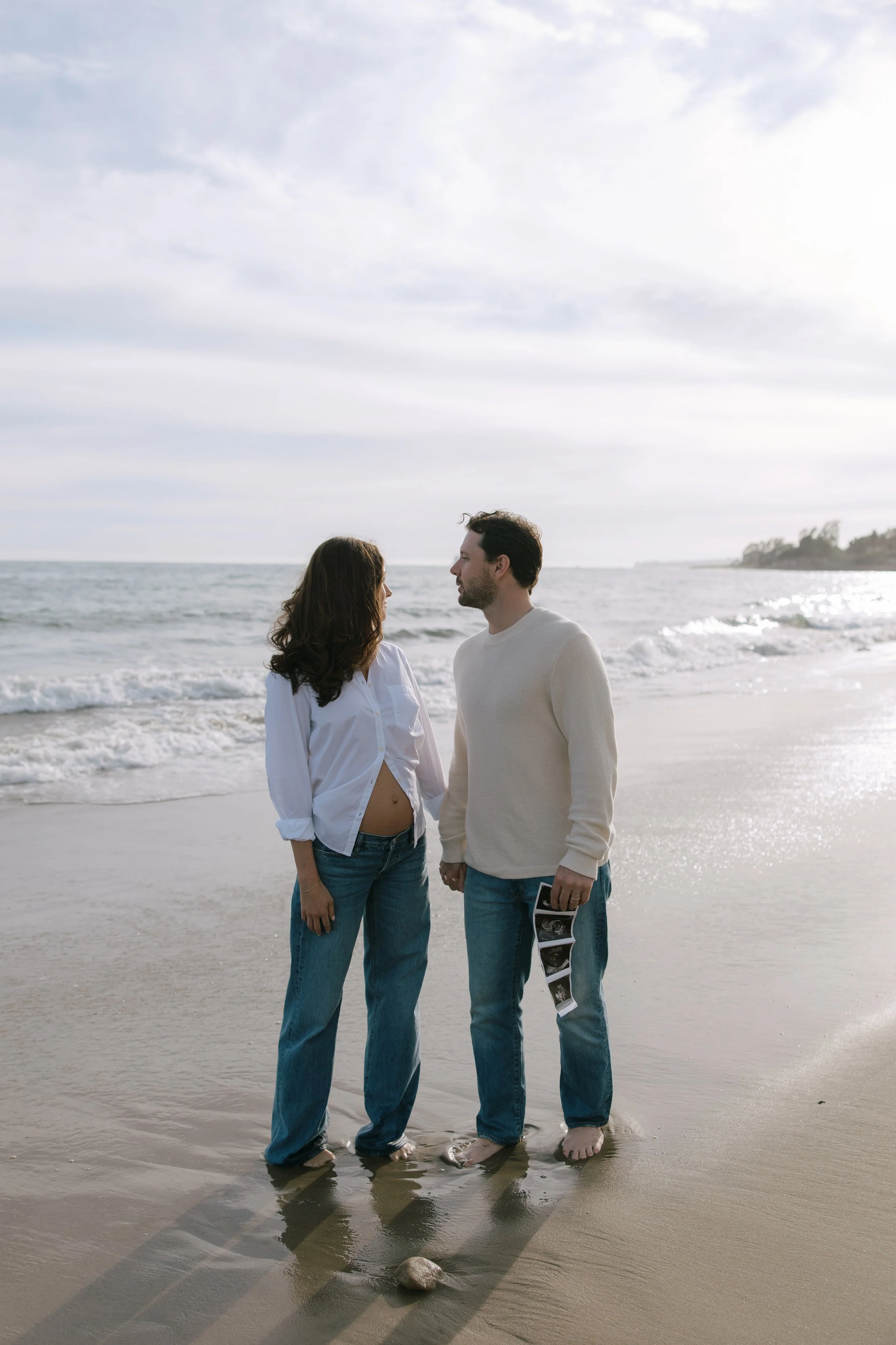 A man and woman standing barefoot on the beach, holding hands, with the ocean in the background, and holding ultrasound pictures.
