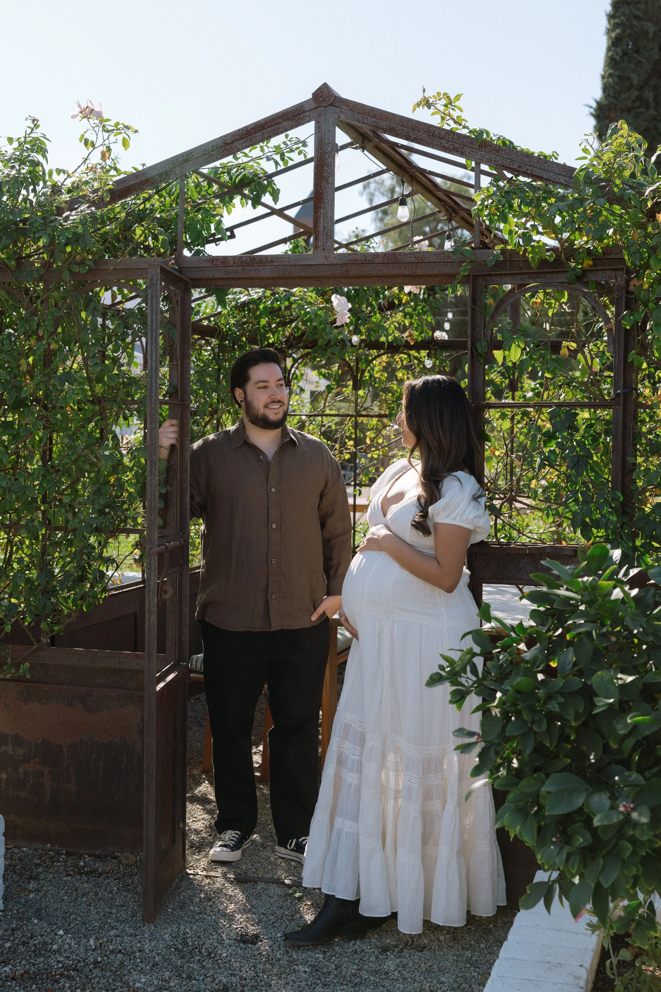 A man and a pregnant woman stand outside in a garden, talking to each other under a rusted metal archway with greenery and flowers around them.