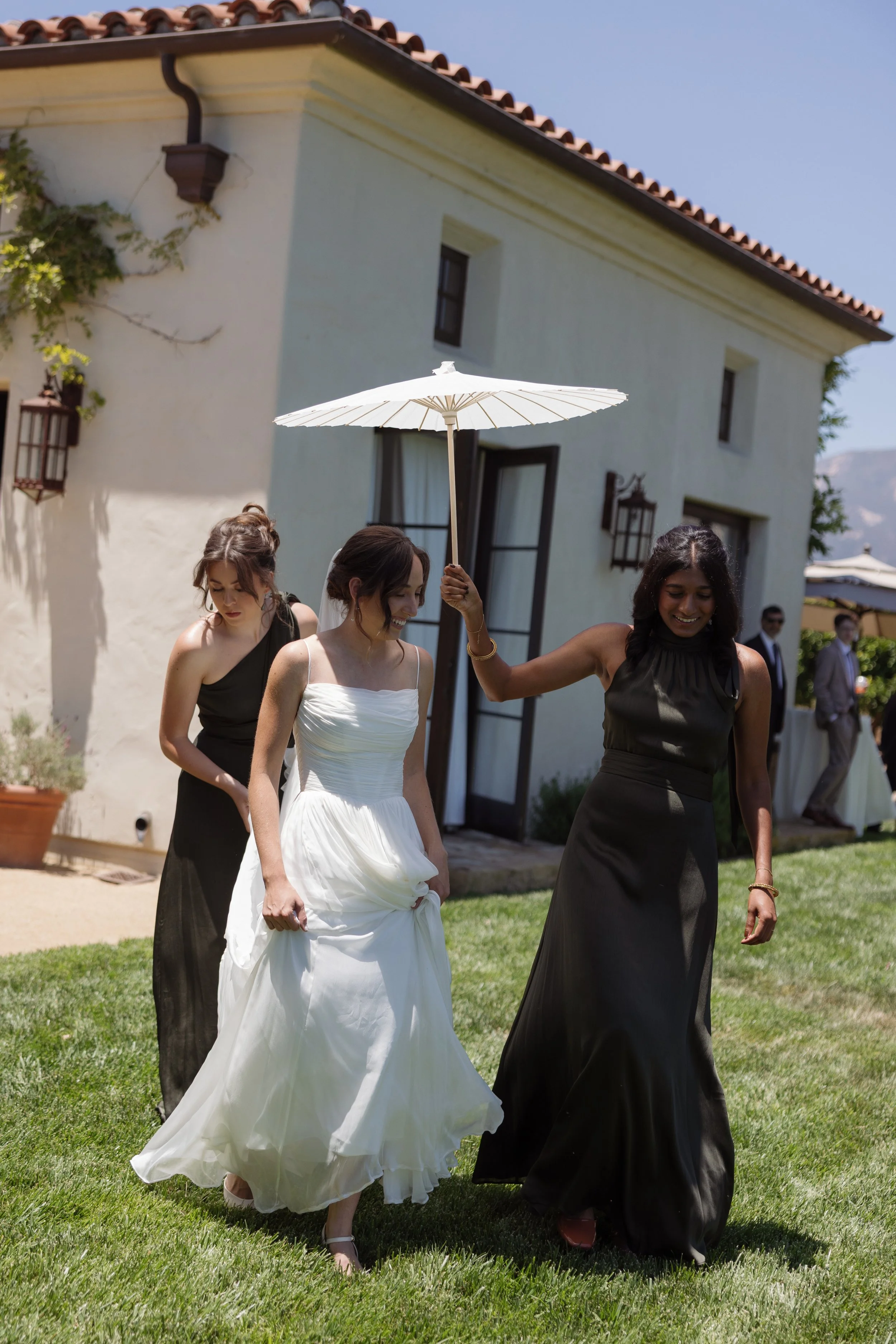 Three women in formal dresses walk on a green lawn outside a white building with a red-tiled roof. One woman holds a white umbrella above her head.