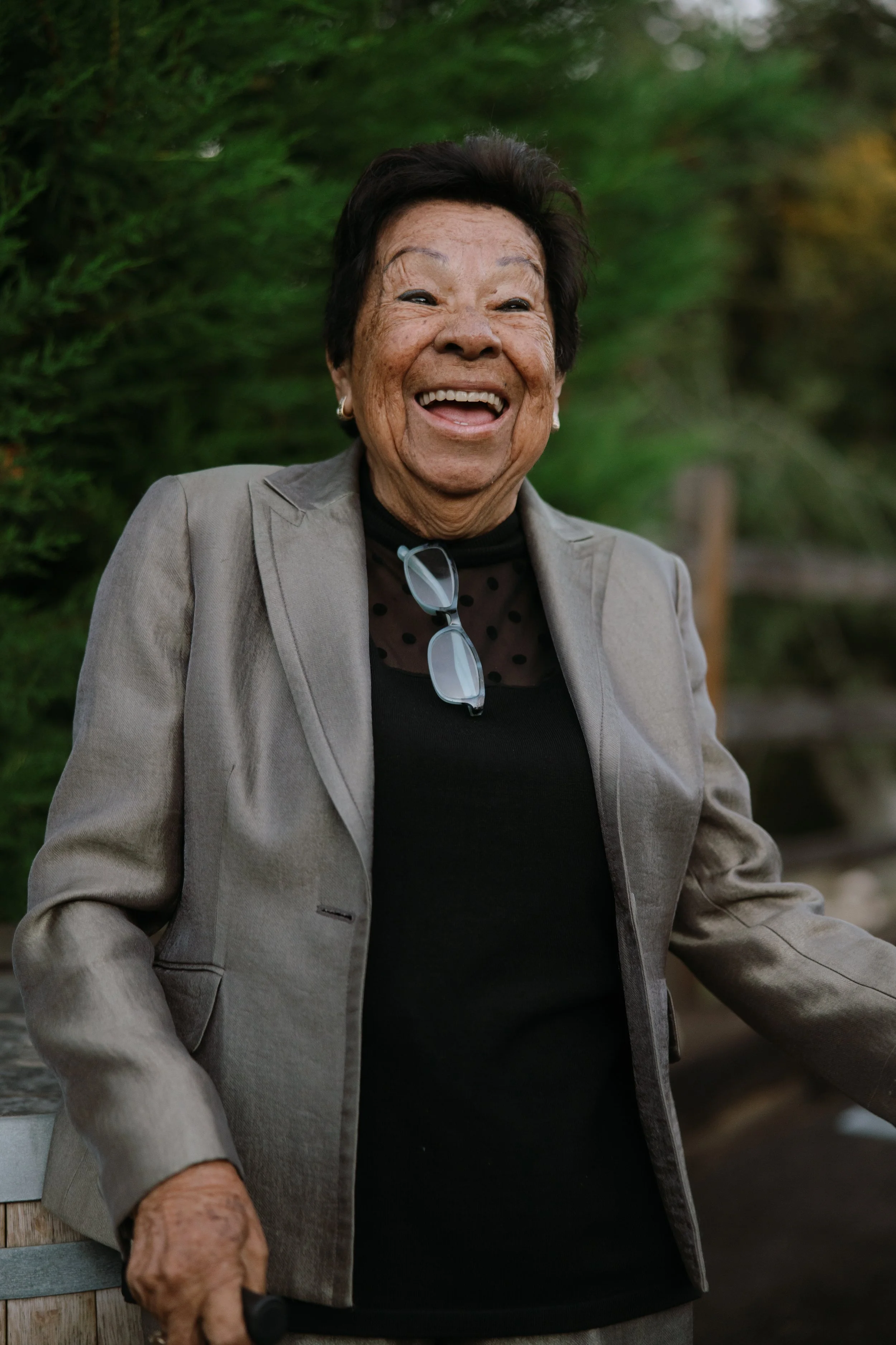 A smiling elderly woman with short dark hair, wearing a beige blazer, black top, with glasses hanging from her neck, standing outdoors in front of green foliage.