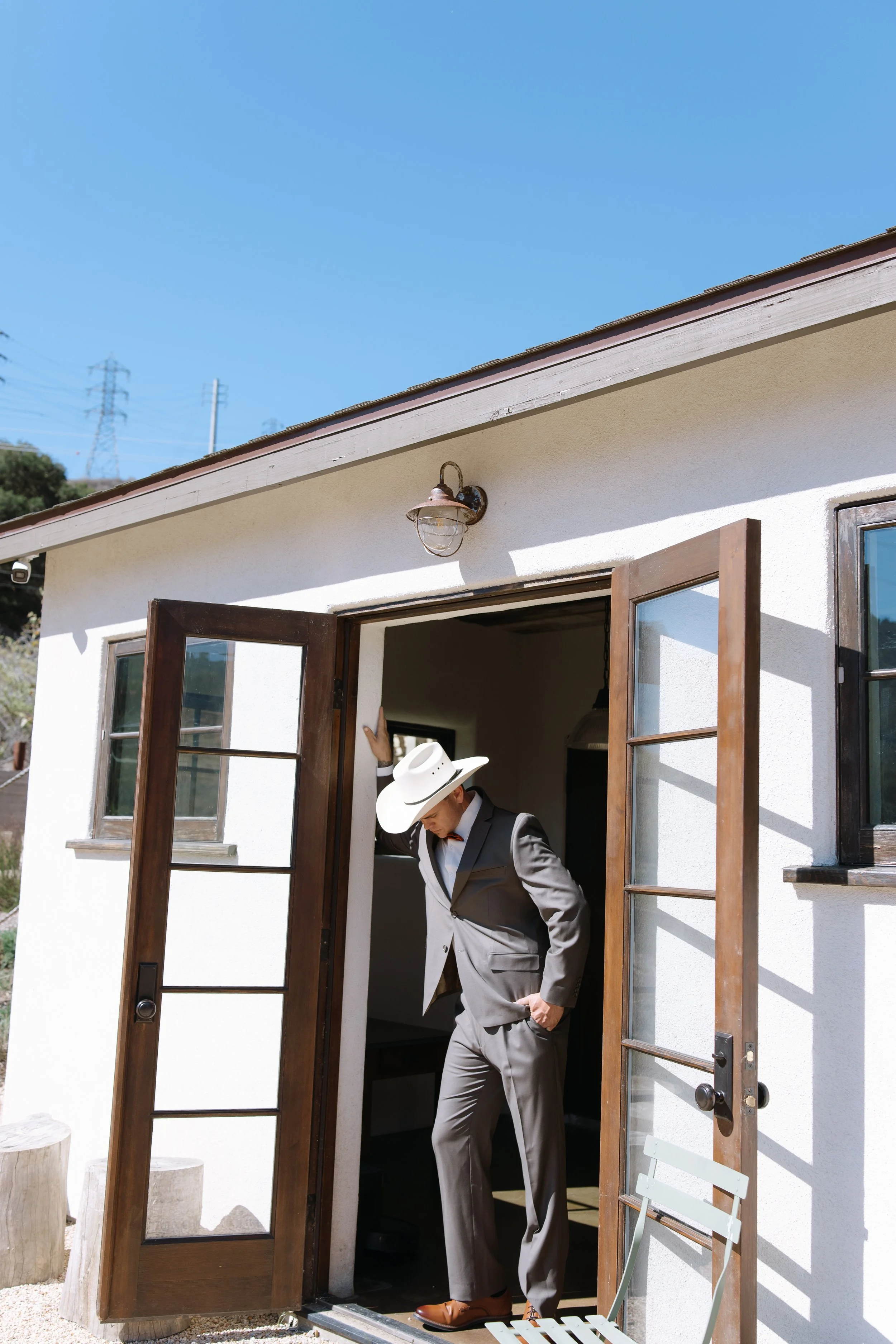 A man in a tan suit and a white cowboy hat is standing in the doorway of a white house with wooden framed glass doors, holding the door open. The house exterior is white with a small outdoor light mounted above the doorway. Outside, there is a light-