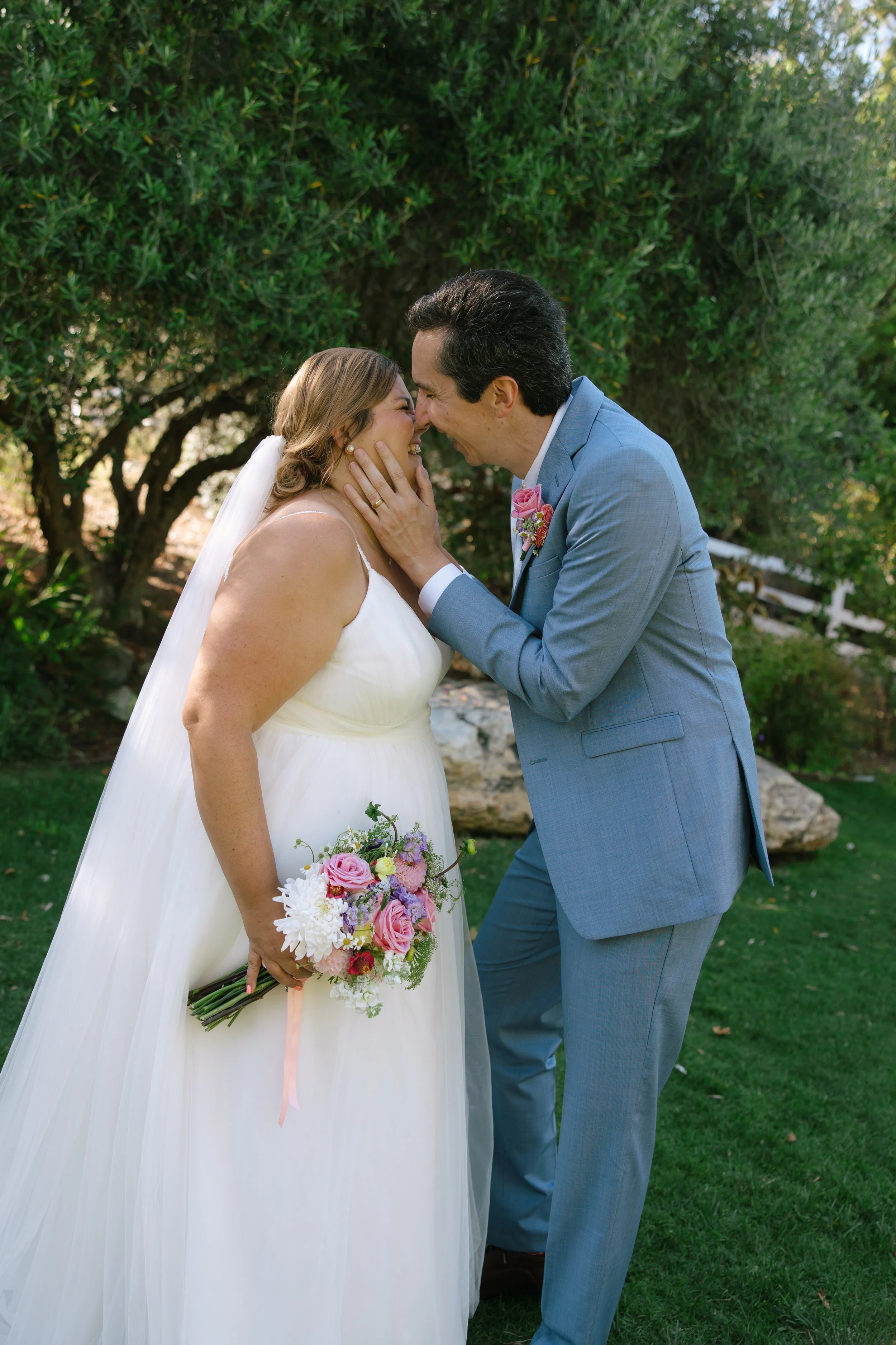 Couple on wedding day with outdoors background of trees and grass, woman in white wedding dress holding a bouquet of pink and purple flowers, man in light blue suit, leaning nose to nose, smiling.