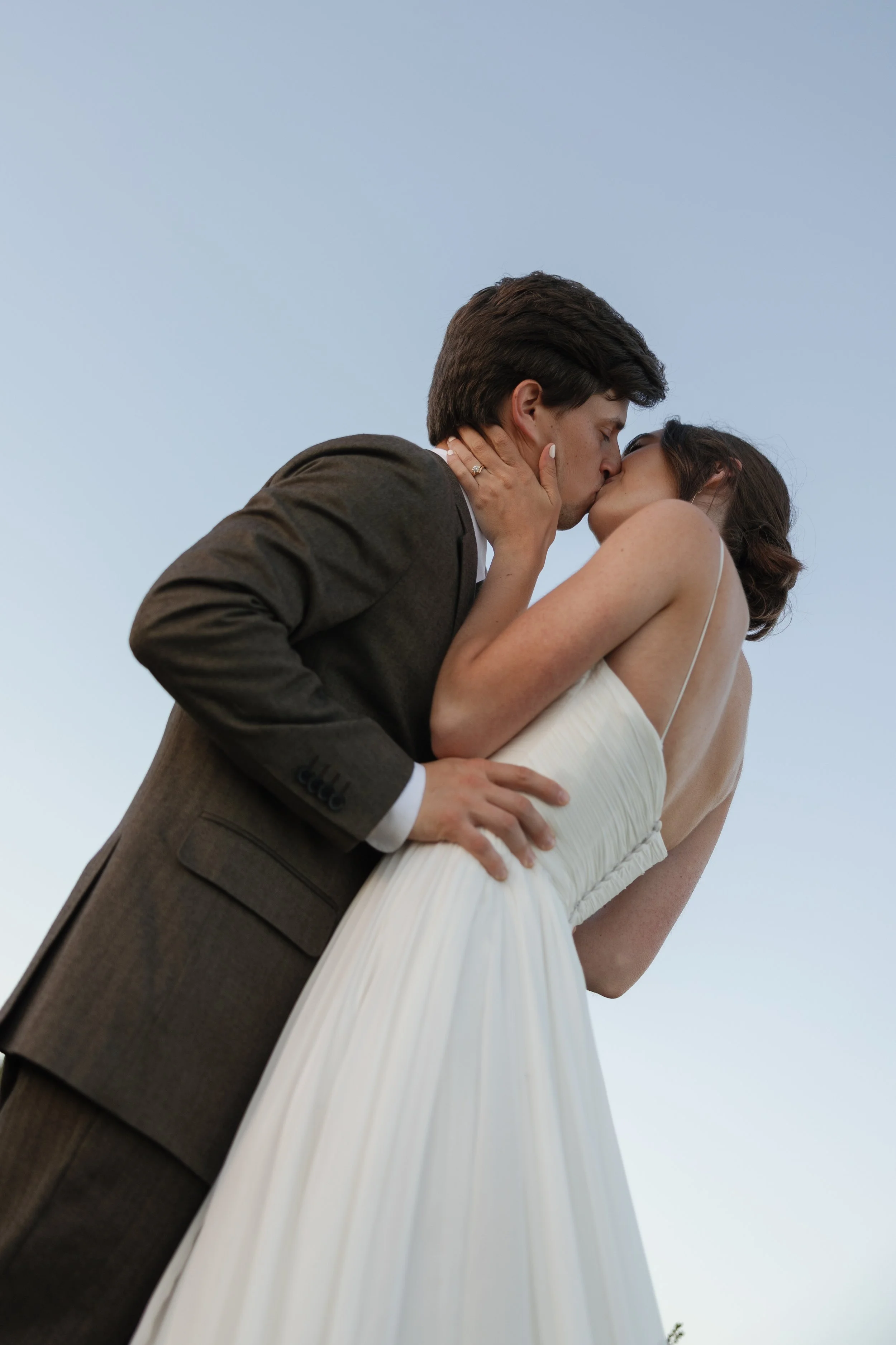 A man and woman share a kiss outdoors against a pale blue sky. The woman is wearing a white dress and the man is dressed in a suit.