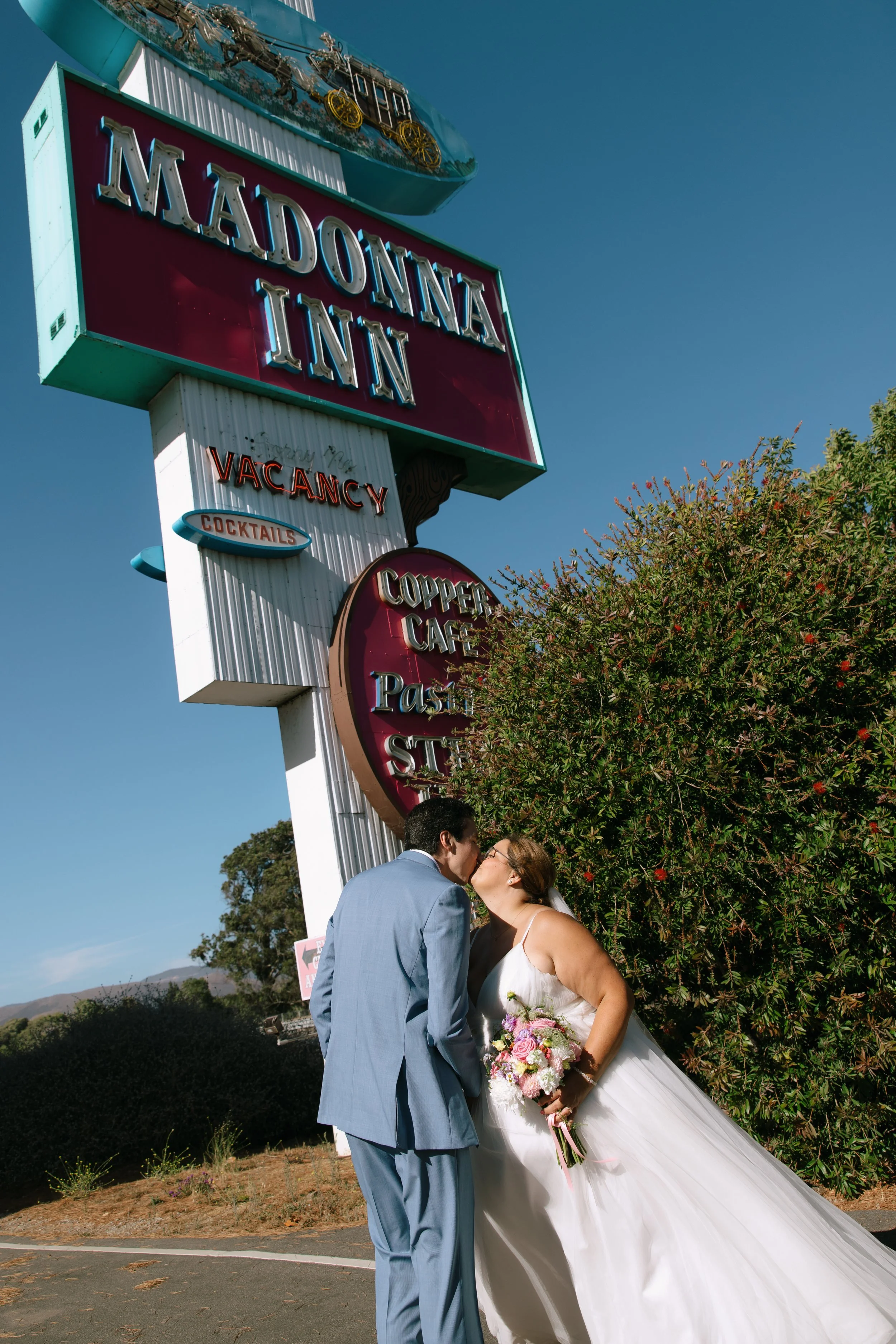 A newlywed couple sharing a kiss outdoors under a blue sky, with a wedding sign in the background and a bride holding a bouquet of flowers.