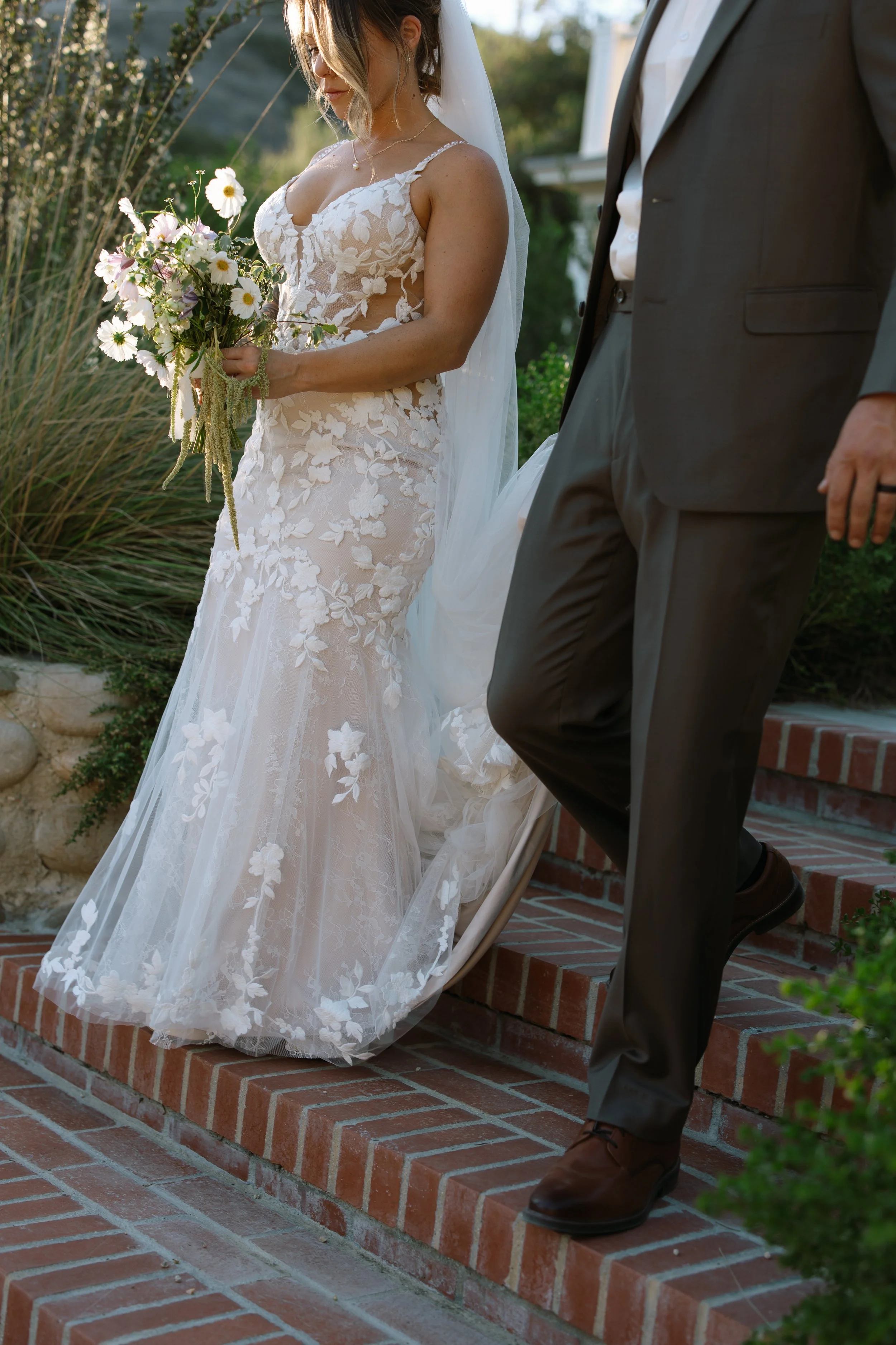 A bride in a white lace wedding gown holding a bouquet of flowers stands on brick steps with a groom in a dark suit. The photo is taken outdoors with greenery in the background.