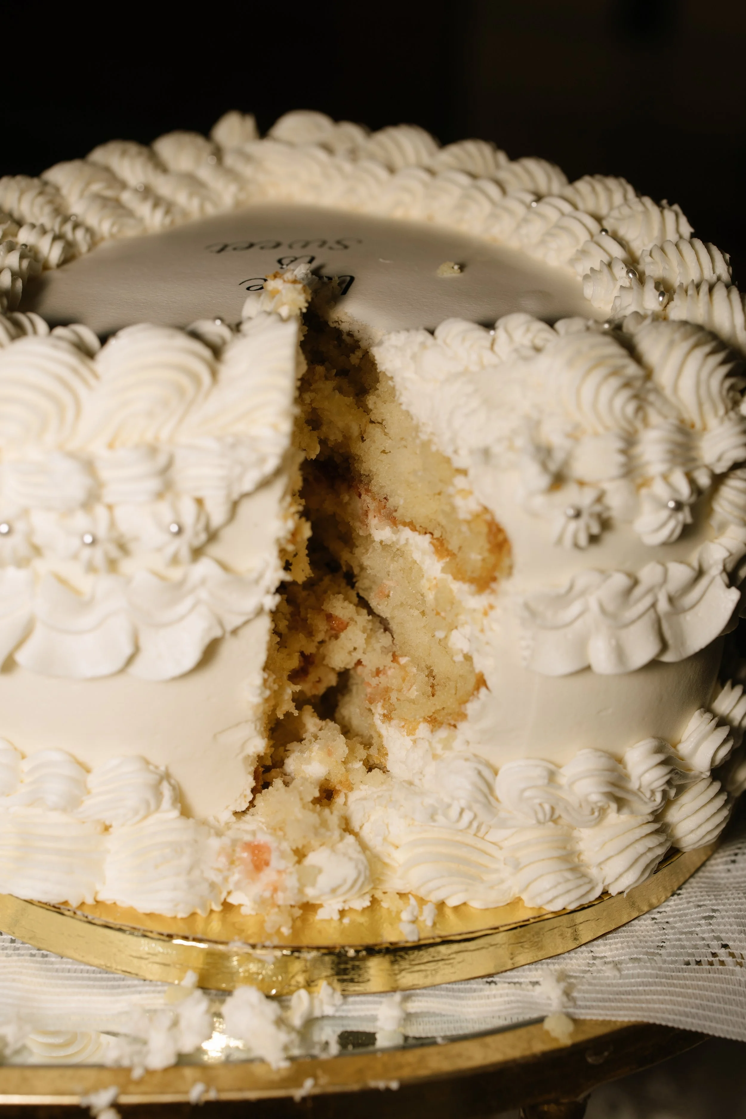 A white, frosted cake with decorative piped icing and silver edible beads, with a slice removed showing a vanilla sponge interior.