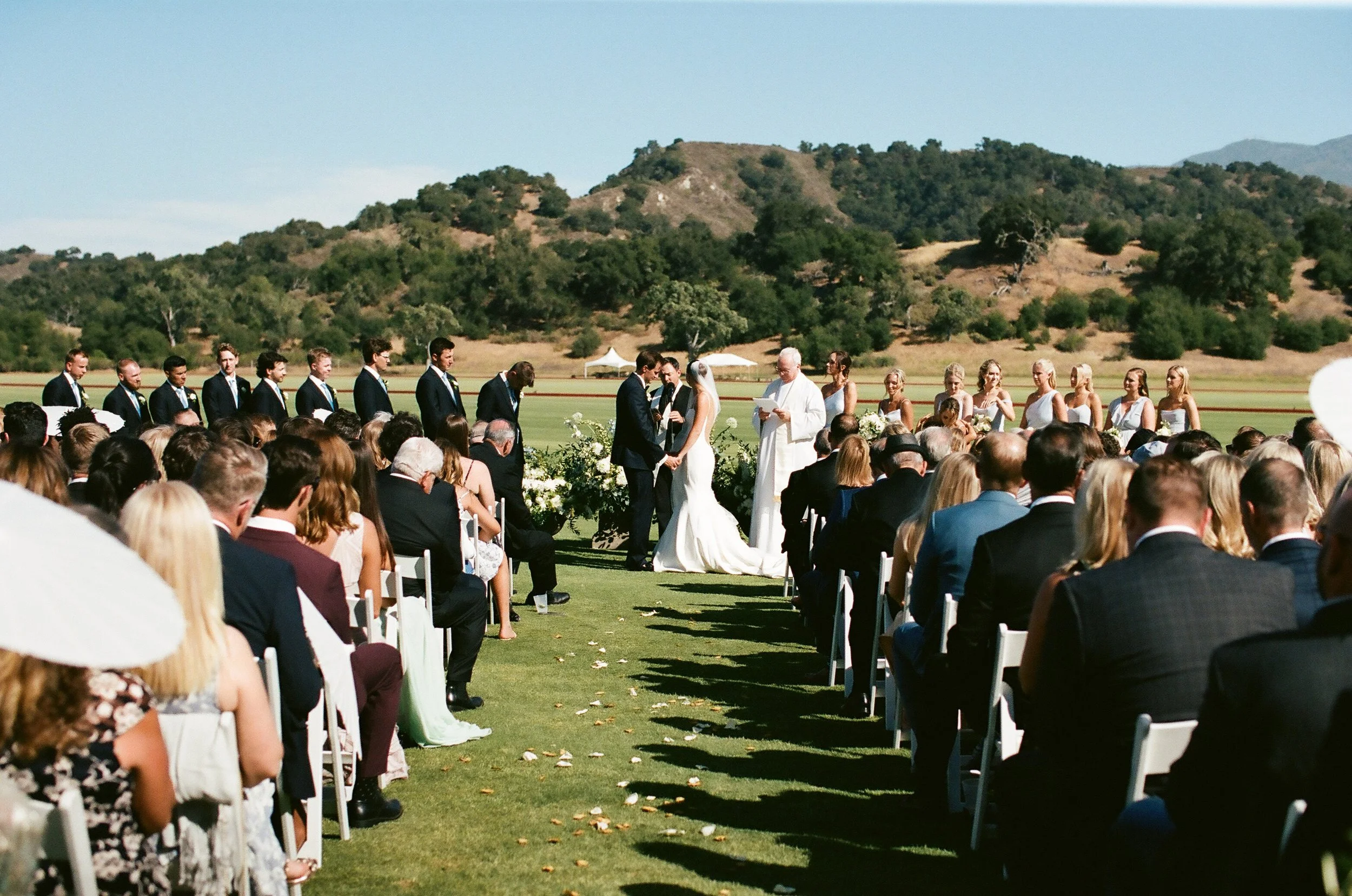 A wedding ceremony outdoors with the bride and groom exchanging vows in front of a priest, surrounded by guests seated on white chairs, with green hills and trees in the background.