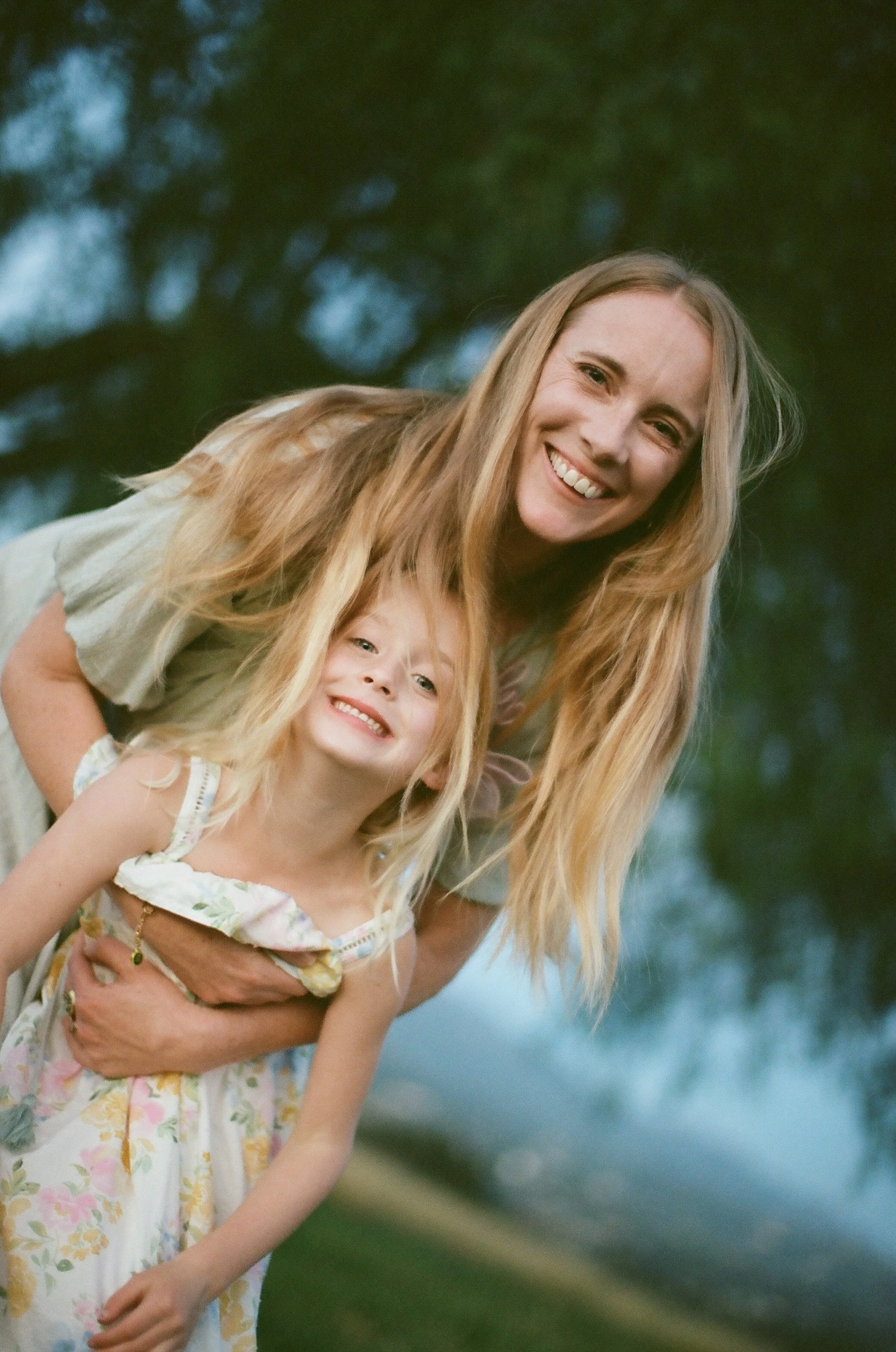 A woman with long blonde hair and a girl with long blonde hair smiling outdoors, with trees and a cloudy sky in the background.