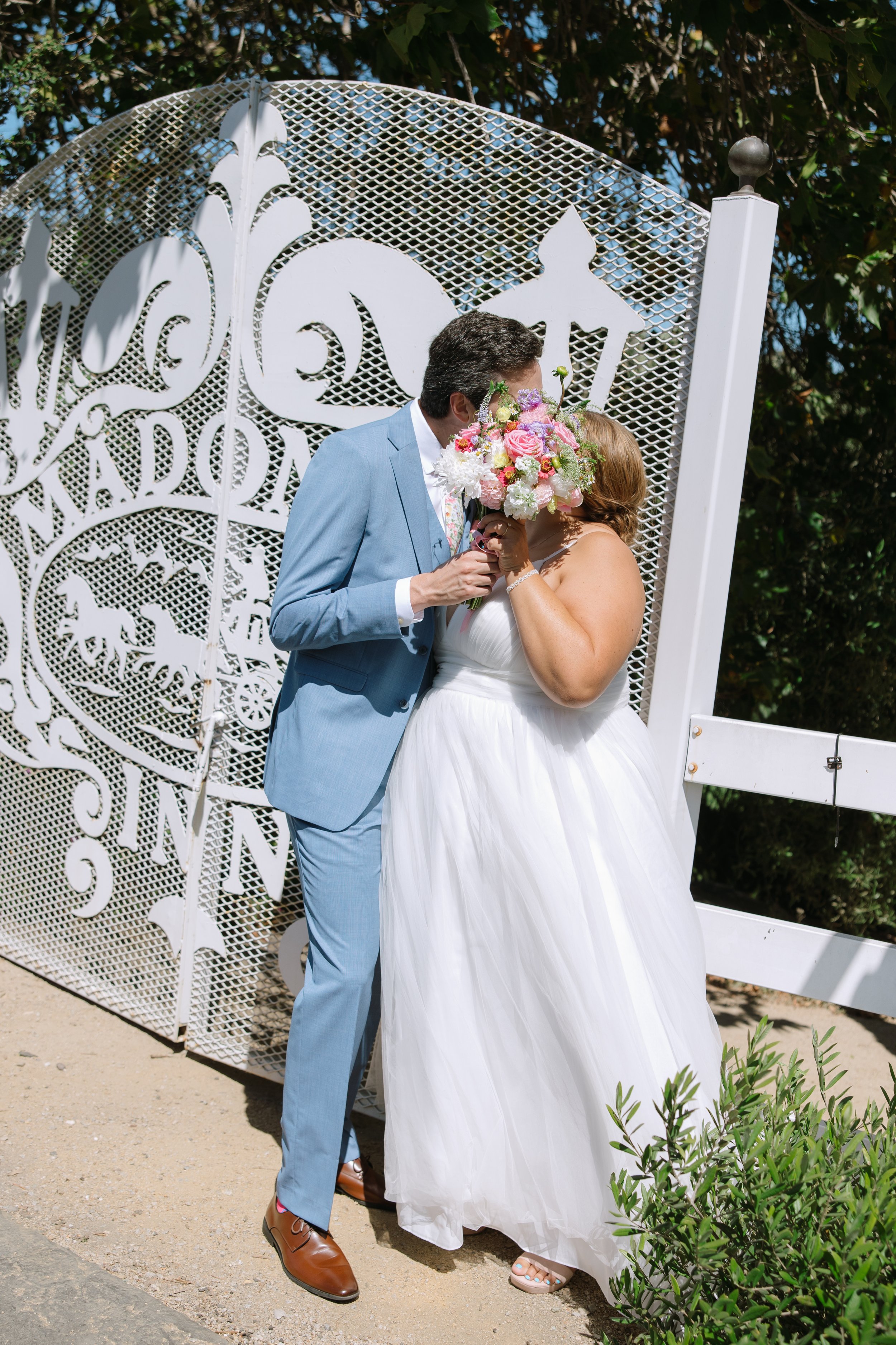 A couple kissing during a wedding ceremony outdoors, with a decorative white metal arch in the background and greenery nearby.