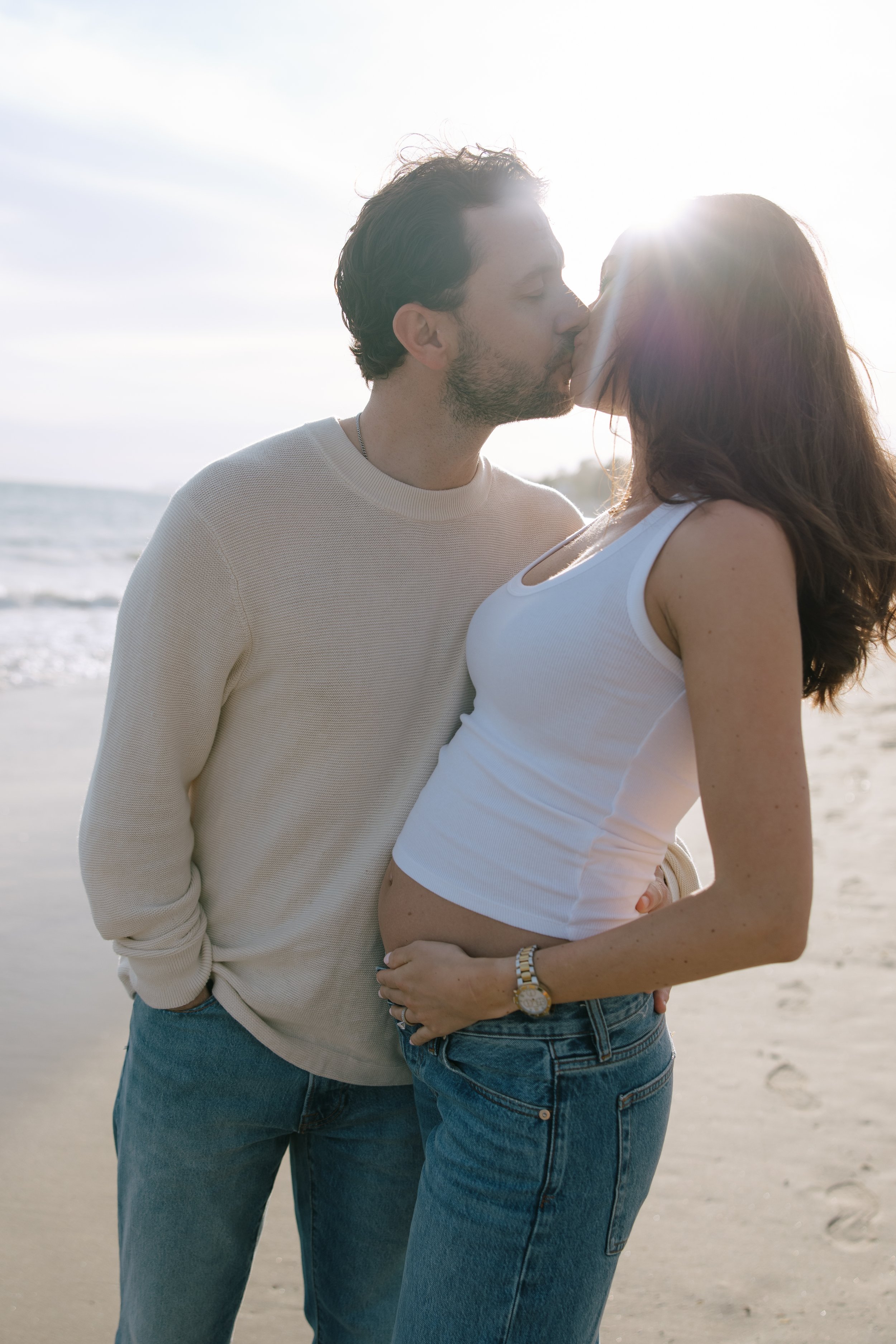 A couple sharing a kiss on the beach during sunset, with the sun shining behind them and the ocean in the background.
