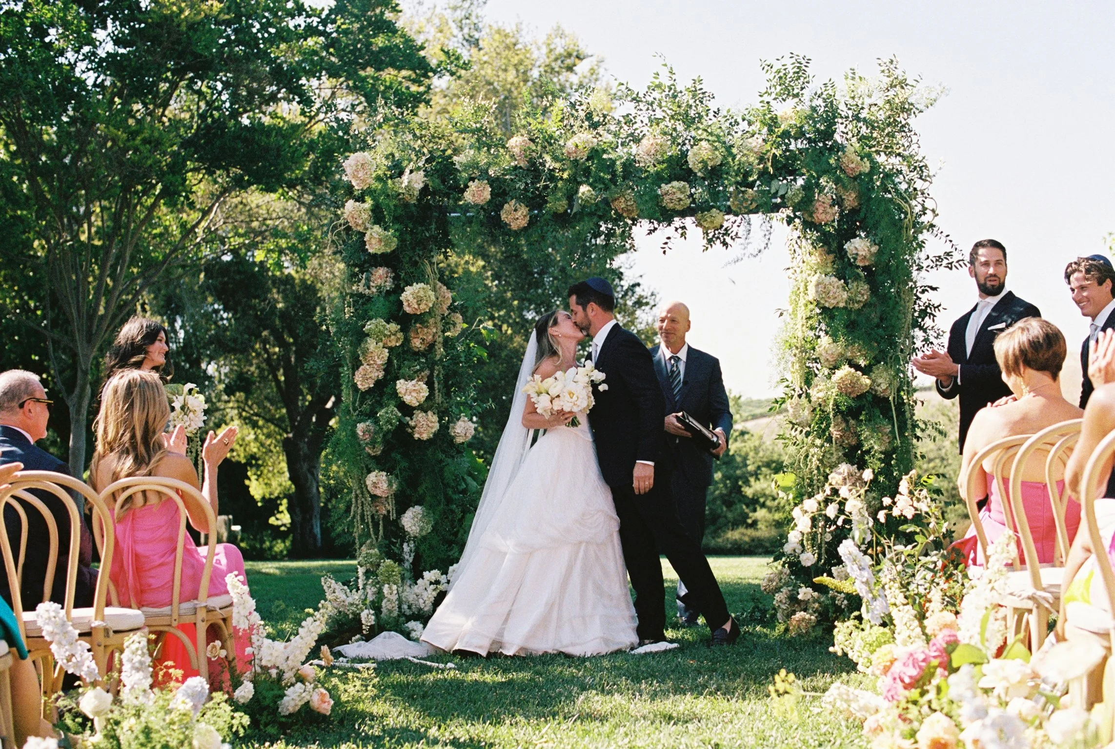 A couple shares a kiss during their outdoor wedding ceremony under a floral arch with guests seated on both sides.