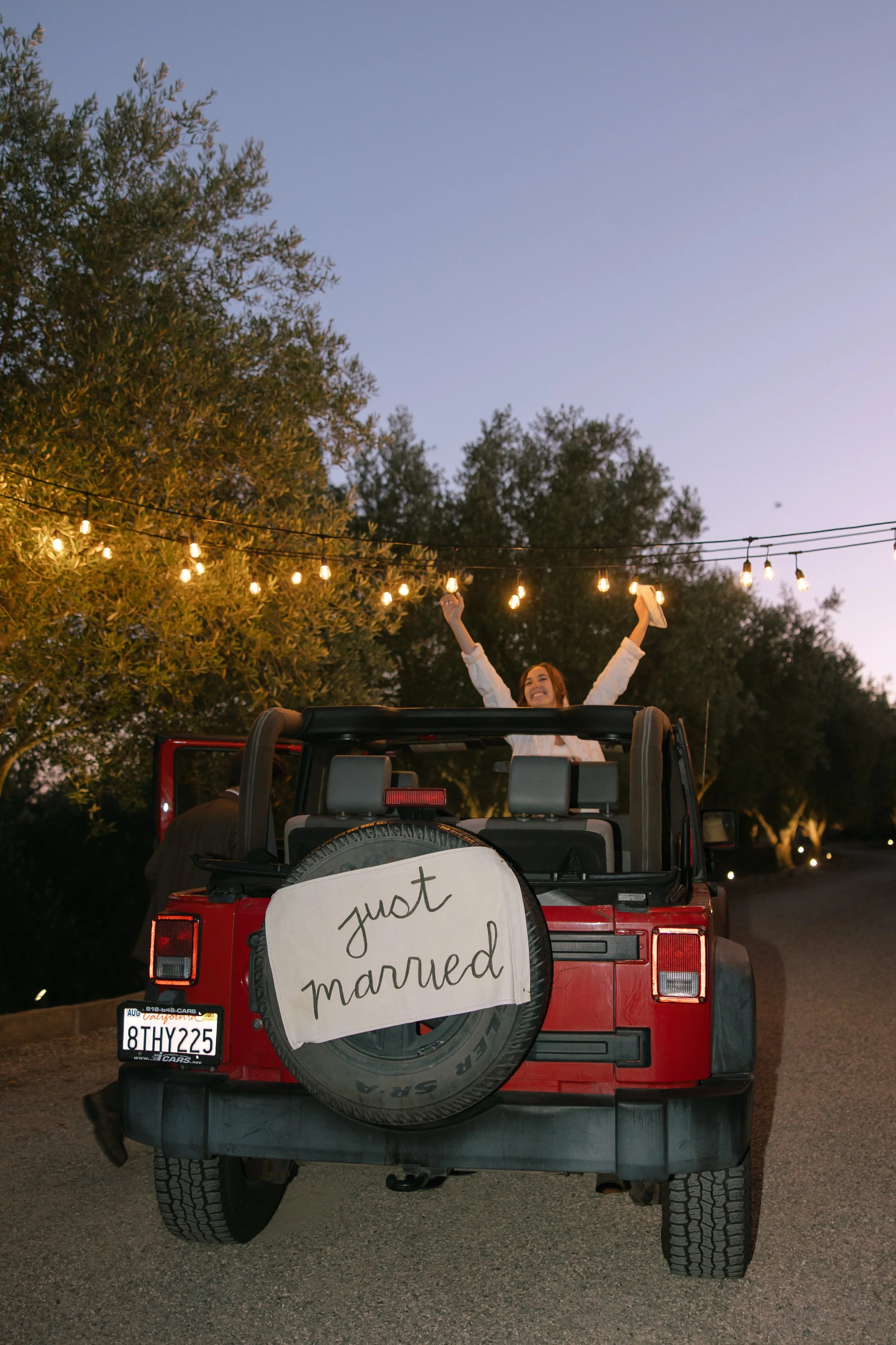 A red Jeep with a 'just married' sign on the back, parked on a street at dusk. A woman is standing in the back of the Jeep with her arms raised in celebration, smiling. String lights are hanging above, and there are trees in the background.