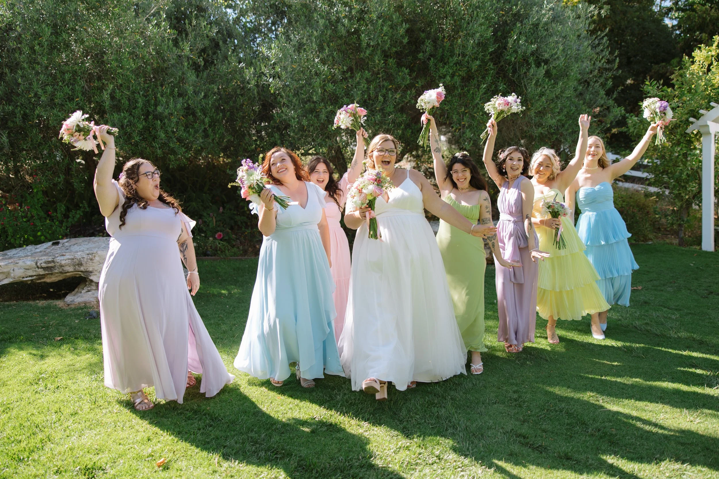 Group of women dressed in pastel-colored dresses celebrating outdoors, holding bouquets of flowers.