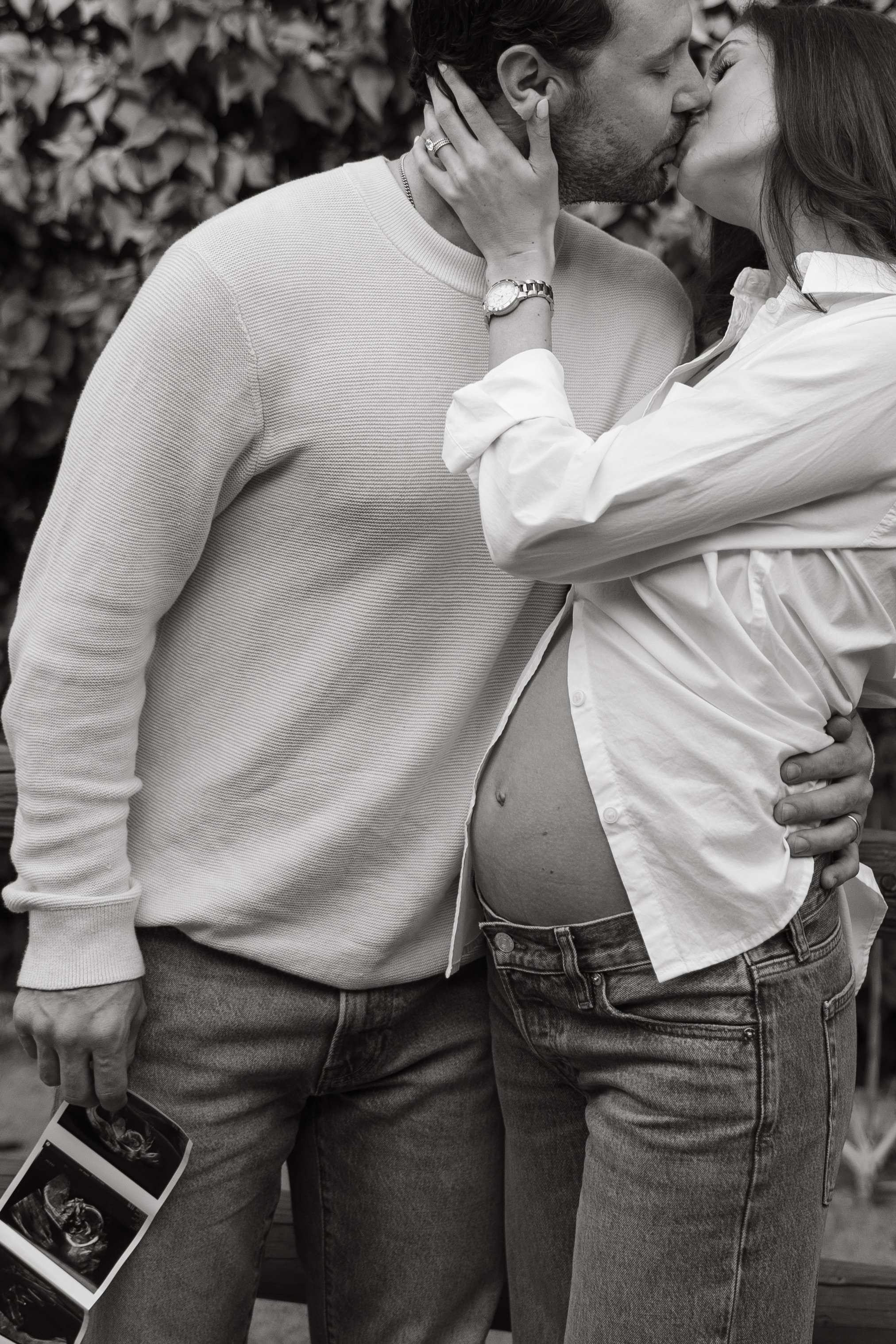 A couple sharing a kiss, with the woman pregnant, outdoors with leafy background.