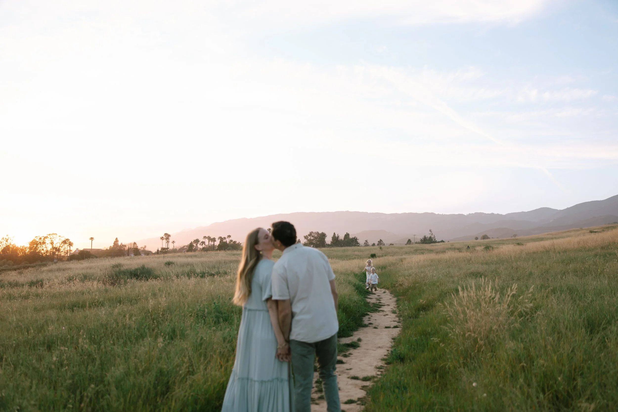 A couple kissing while walking through a grassy field at sunset, with two children walking behind them on a dirt path, mountains in the distance and a cloudy sky.