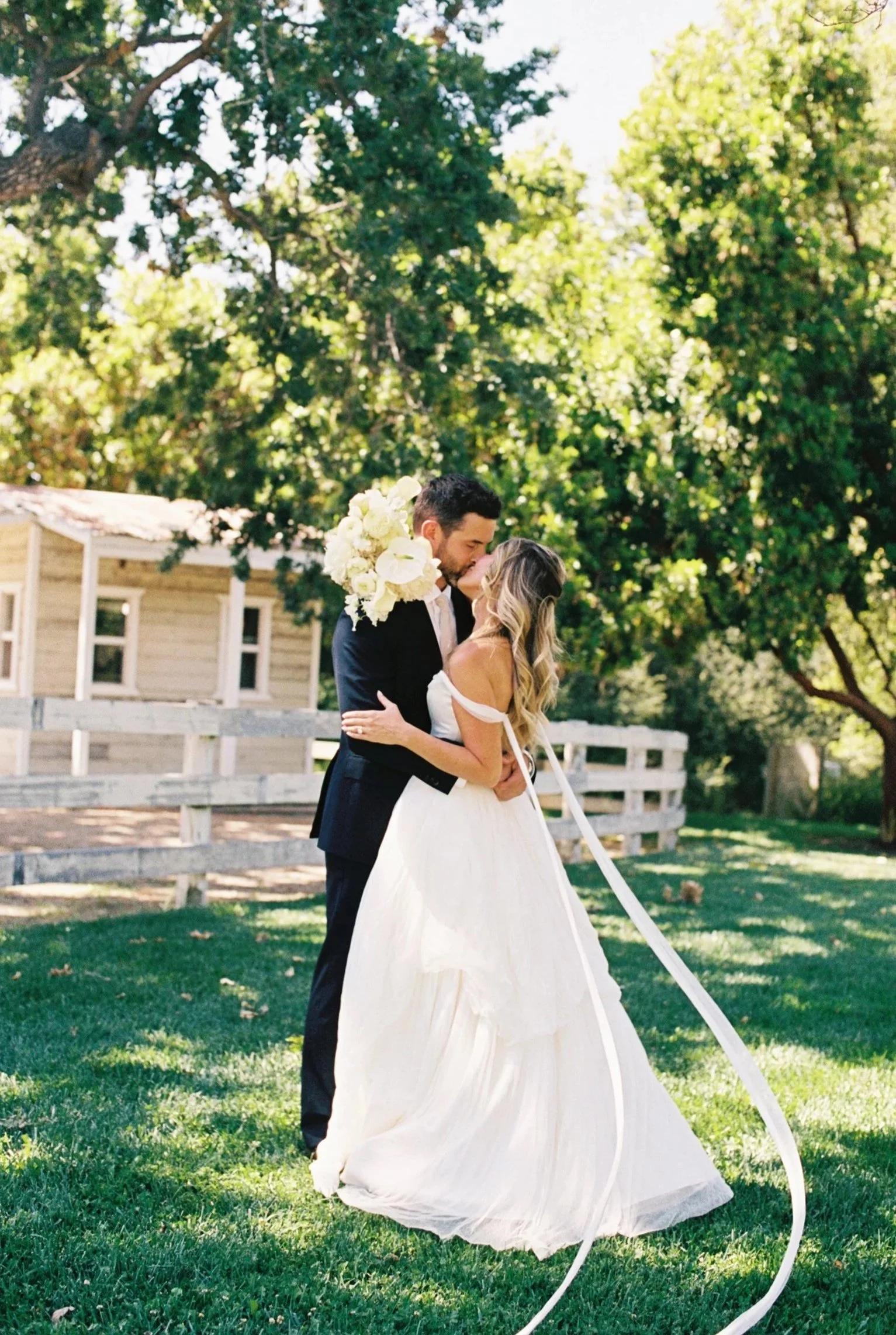 A bride and groom sharing a kiss outdoors during their wedding, with trees and a wooden house in the background.