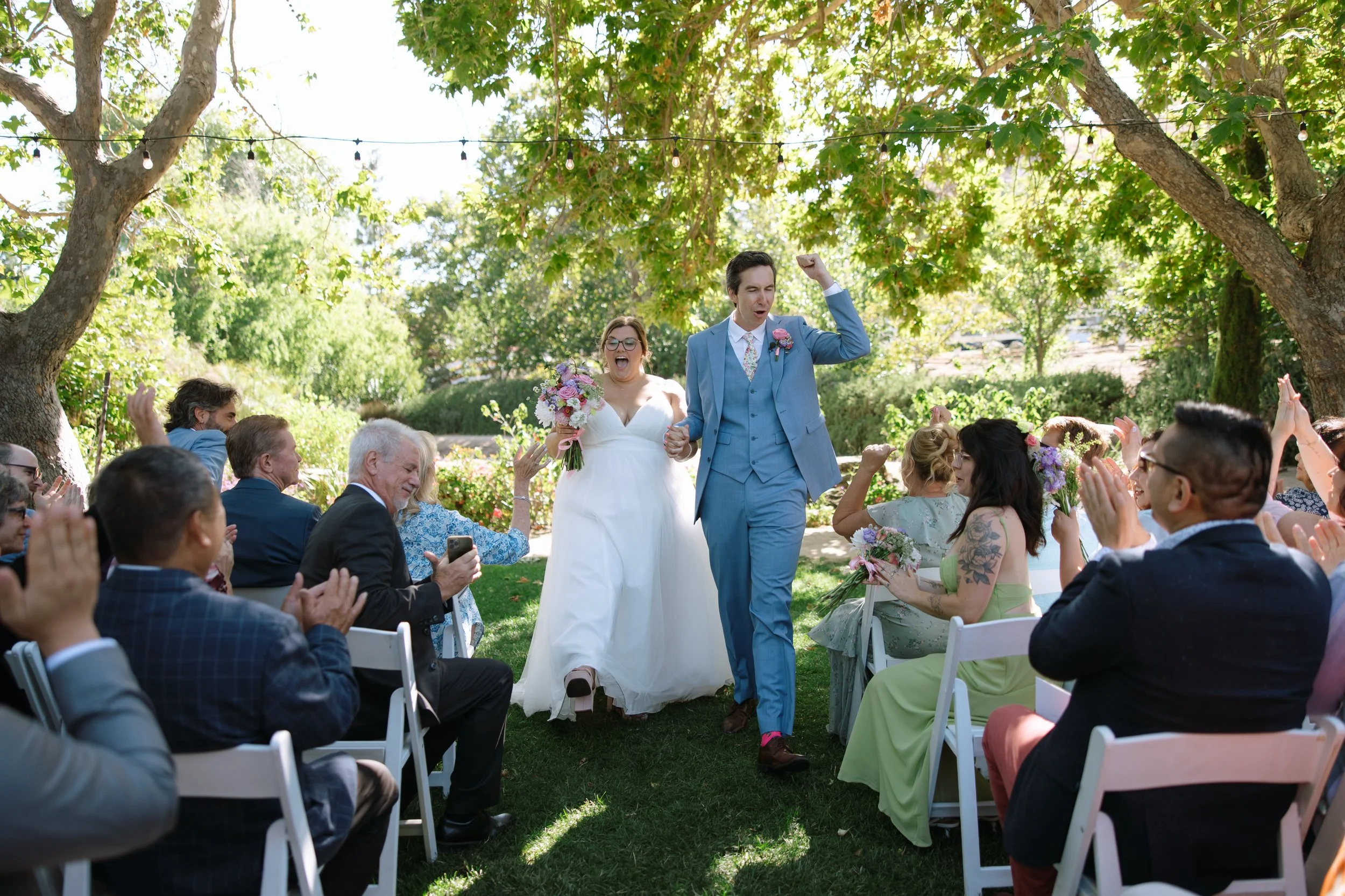 Bride and groom walking down the aisle at an outdoor wedding ceremony, surrounded by seated guests and trees.