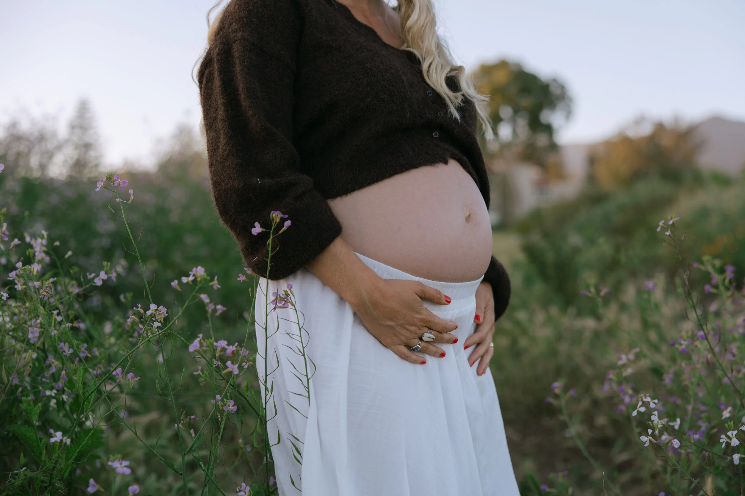 A pregnant woman stands in a field of purple flowers, wearing a black sweater and a white skirt, with her hands resting on her belly.