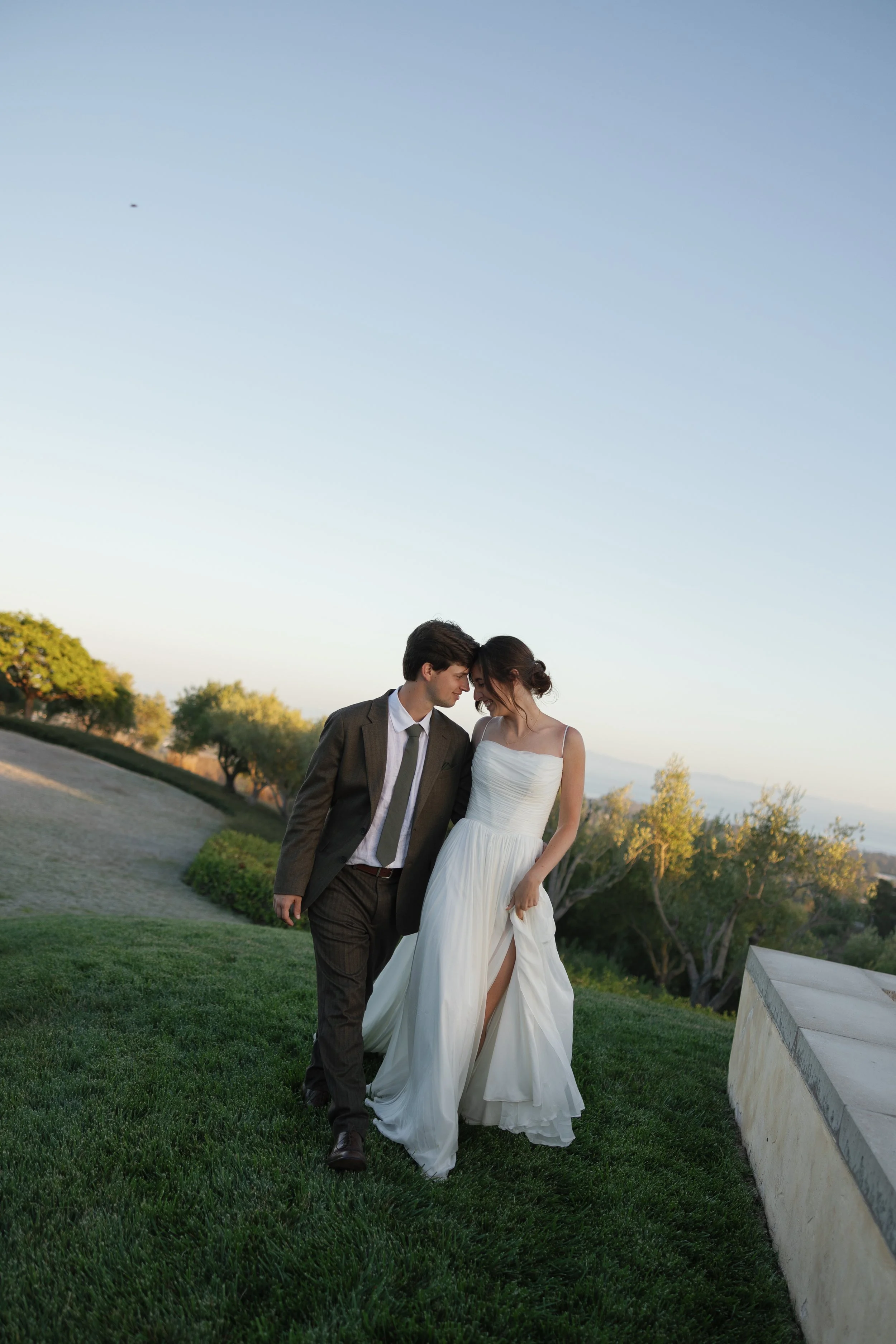 A couple, a man in a brown suit and tie and a woman in a white wedding dress, walk closely together on grass with trees and a blue sky in the background during sunset.
