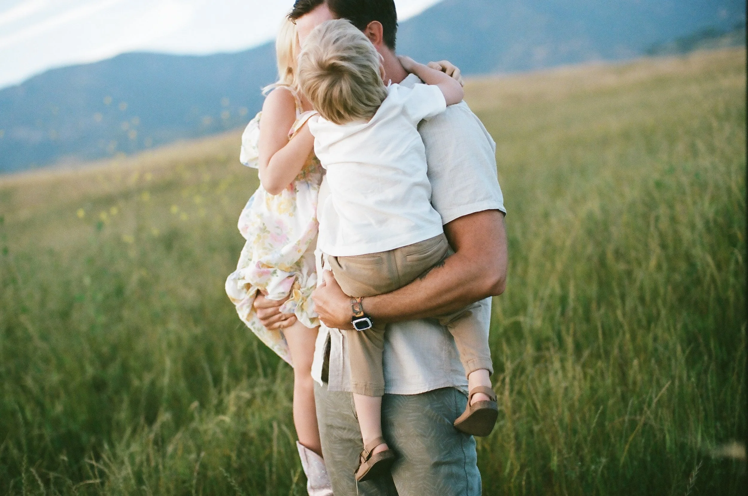 A man holding two children in a grassy field with mountains in the background, embracing each other.