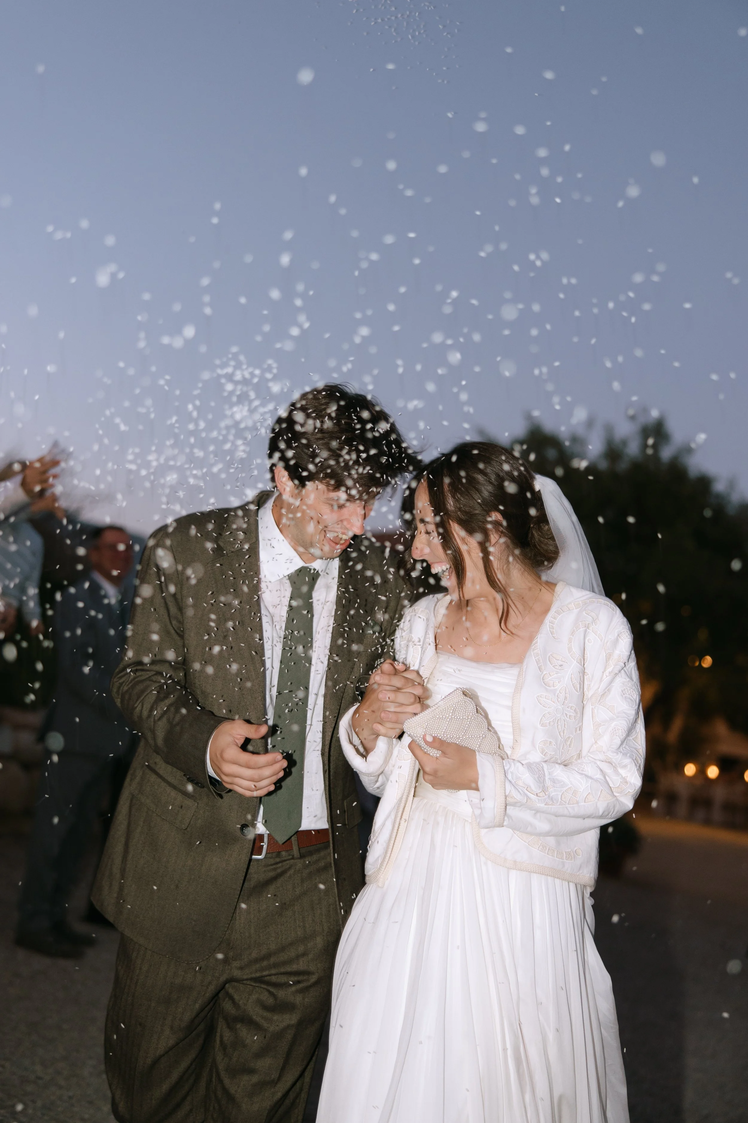 A bride and groom celebrating at their wedding, smiling and holding hands, surrounded by guests throwing rice or confetti into the air during the evening outdoors.