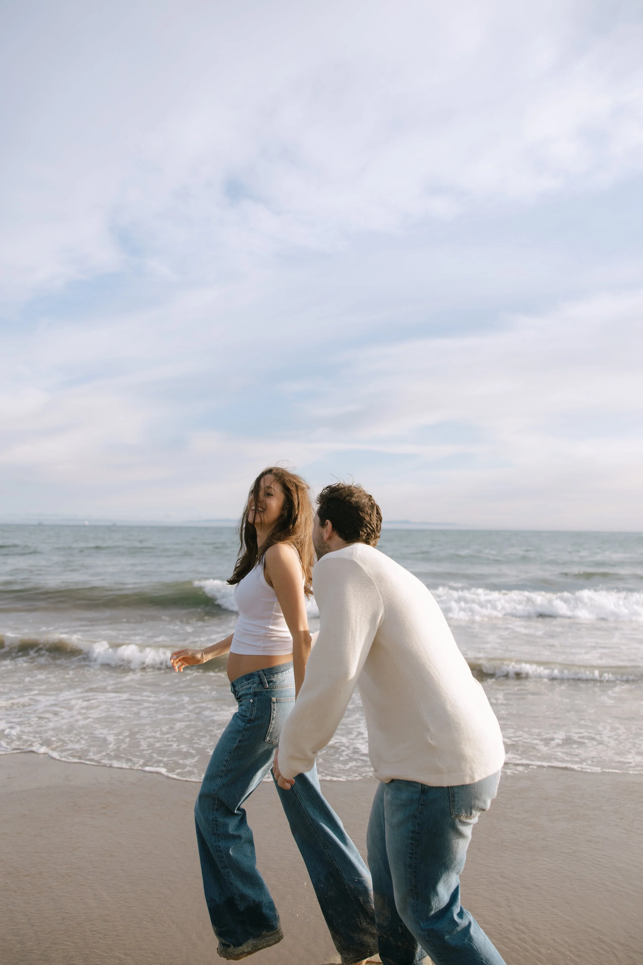 A young woman and a young man walk along the shoreline at the beach, with the ocean waves behind them and a cloudy sky above.