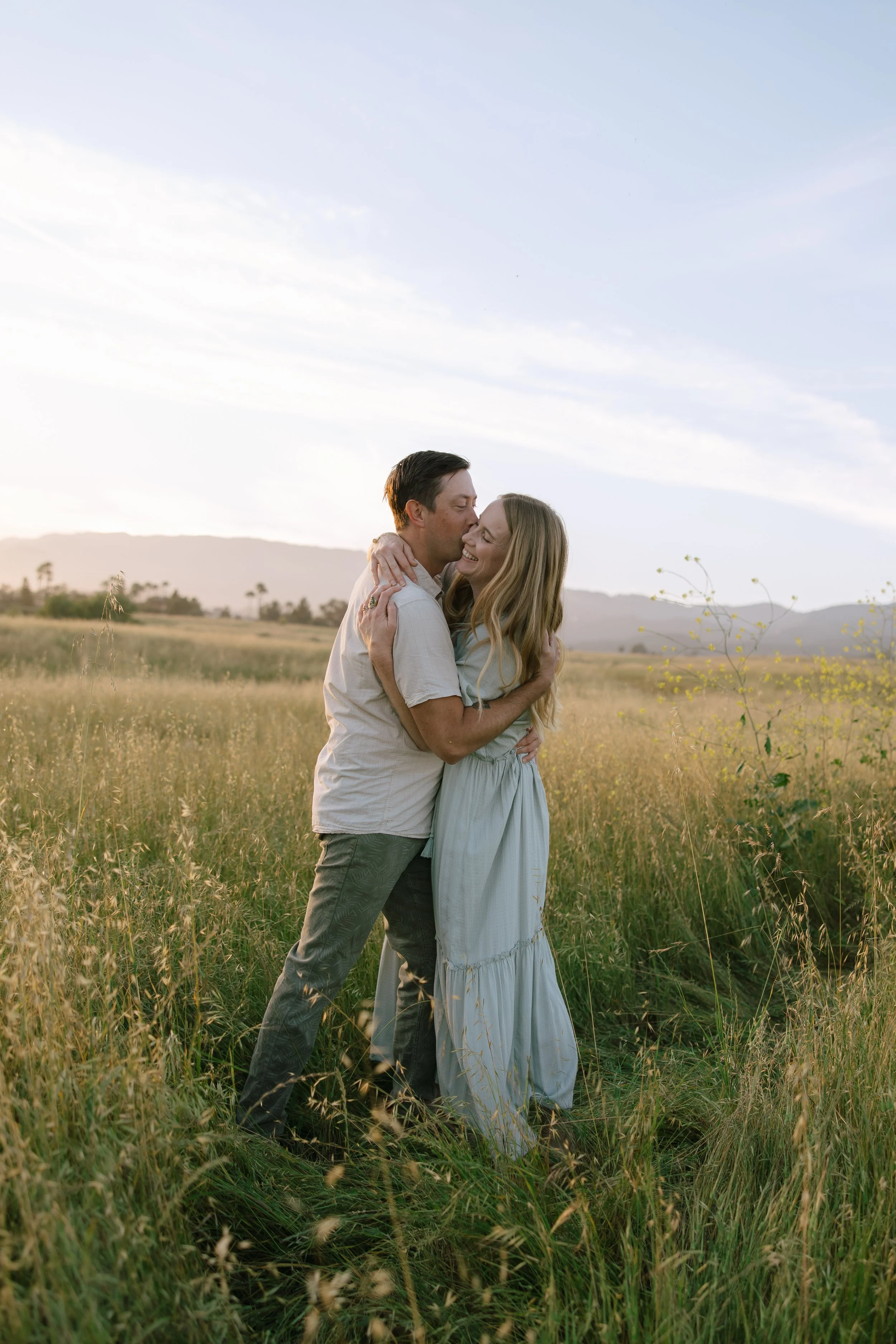 A couple embracing and smiling in a grassy field during sunset. The man is kissing the woman on the forehead, and they are both wearing light-colored, casual clothing.