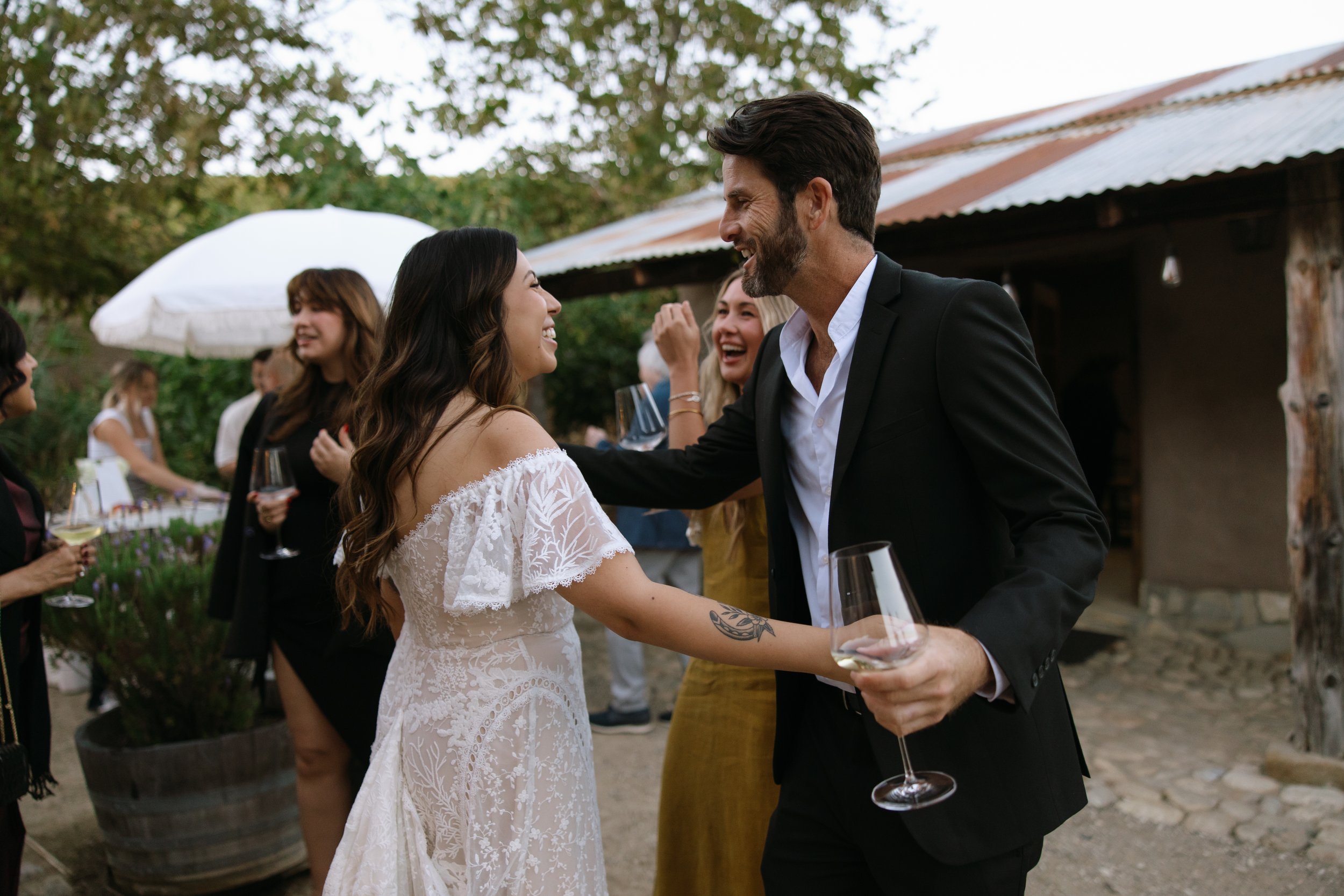 A couple sharing a joyful moment at an outdoor wedding reception, surrounded by friends with drinks in hand, under a large white umbrella with trees and rustic building in the background.