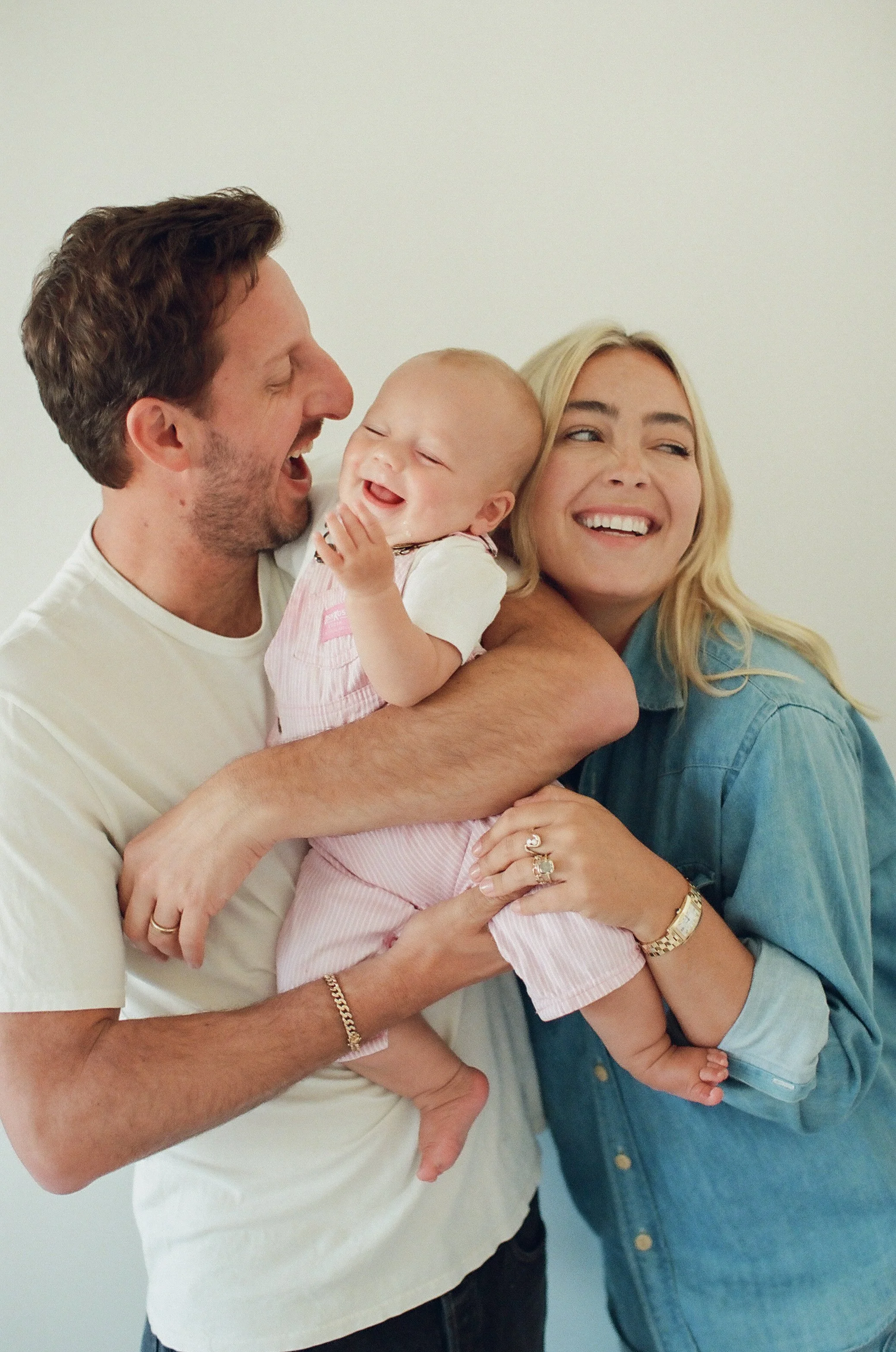 A happy family of a man, woman, and baby laughing and embracing each other against a plain background.