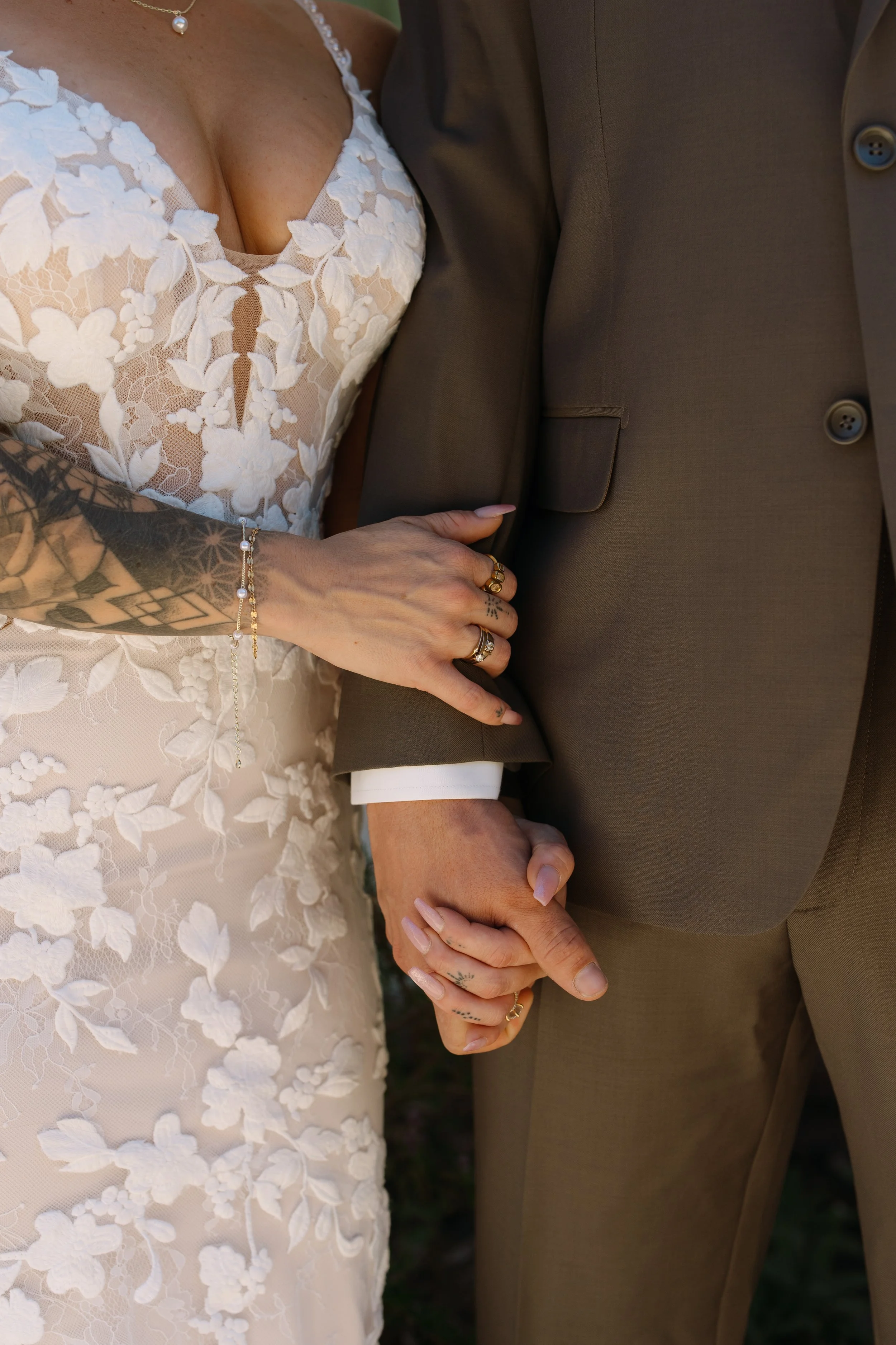 Close-up of a bride and groom holding hands, with the bride wearing a white lace dress with floral appliqués and a pearl necklace, and the groom wearing a brown suit.