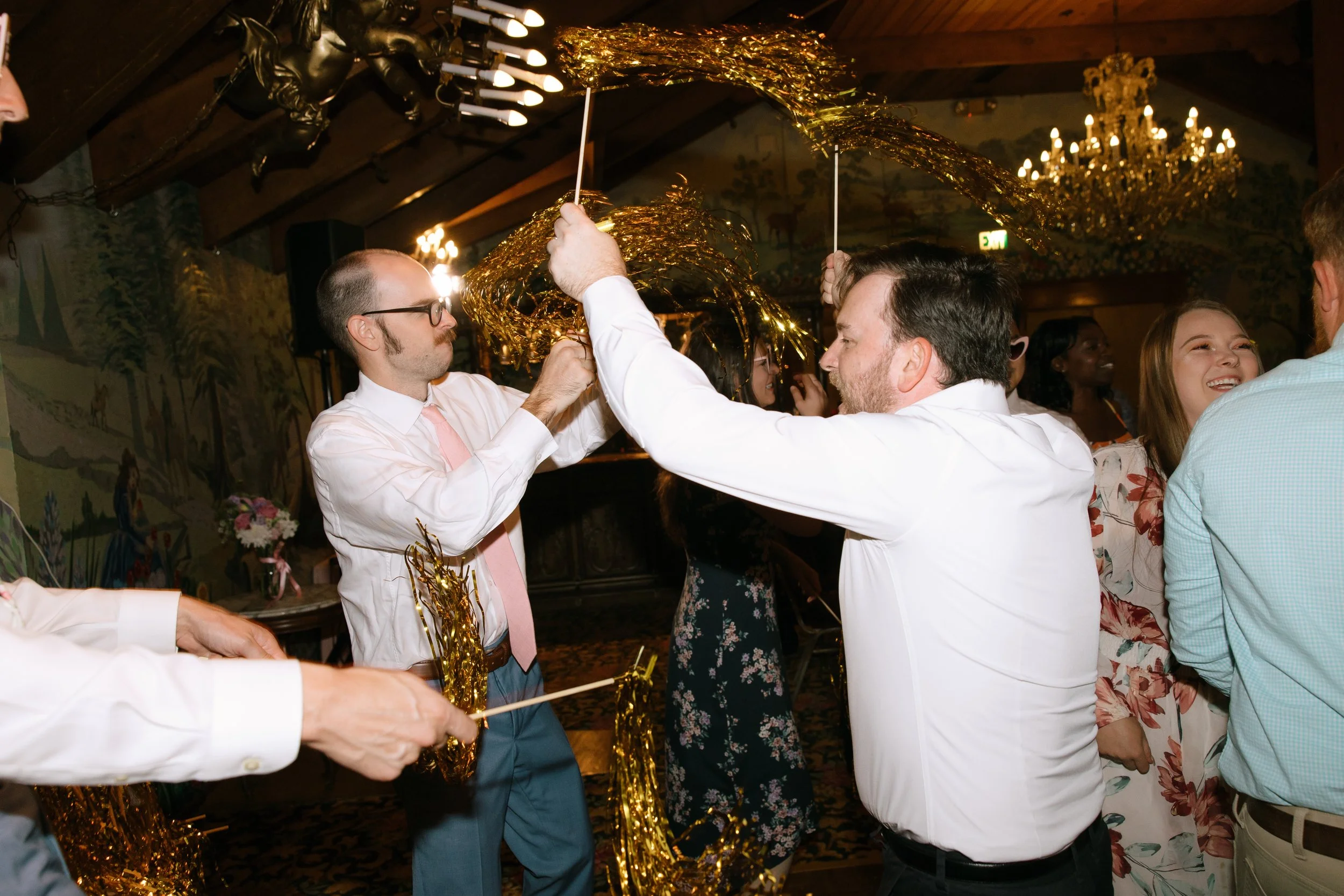 People celebrating at a party, holding up gold tinsel and balloons, with smiles and laughter, in a decorated indoor venue with chandeliers and mural paintings on the wall.