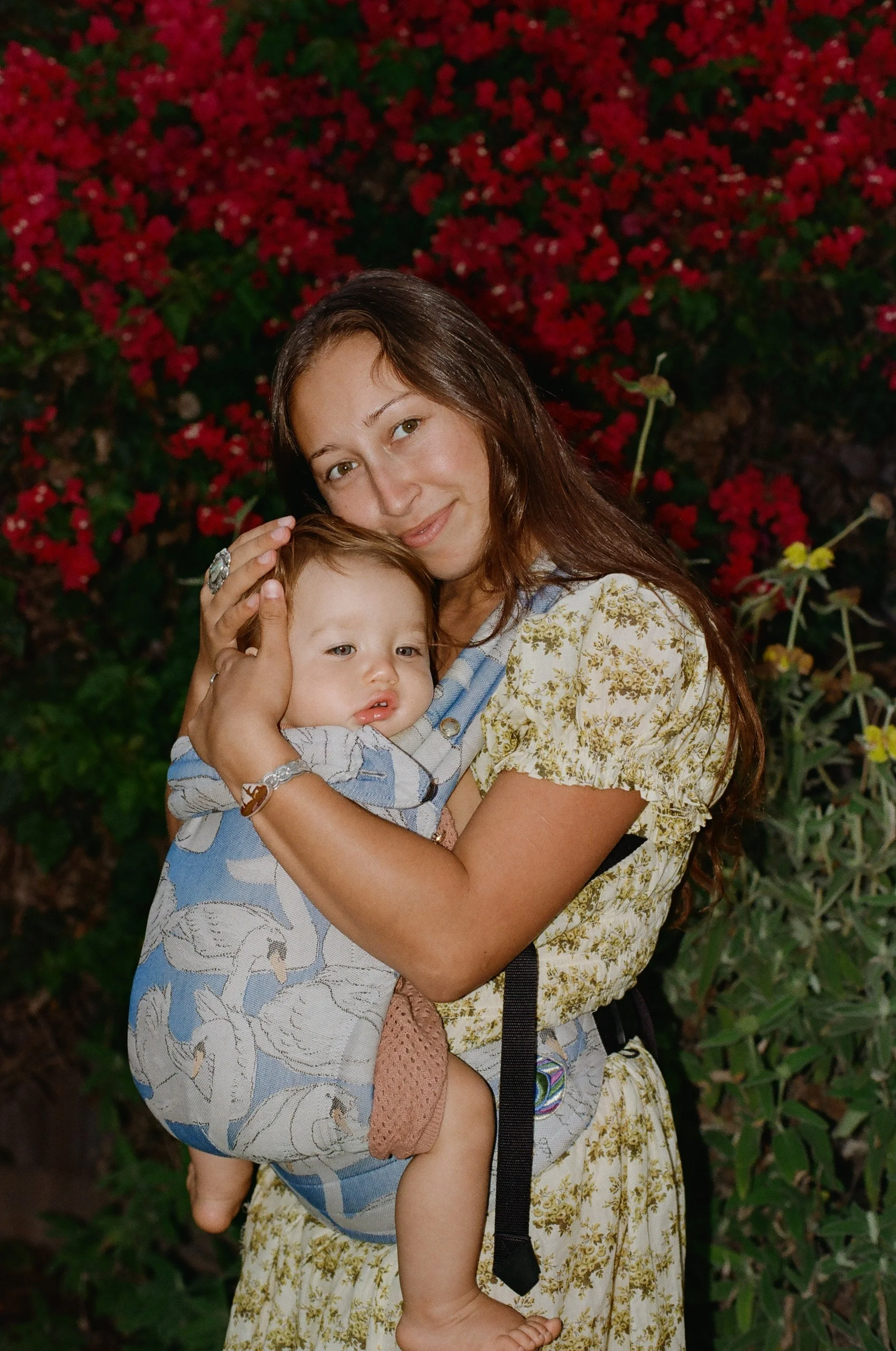 A woman with long brown hair smiling and holding a young boy with light brown hair, both standing in front of a background of pink and red flowers and greenery.