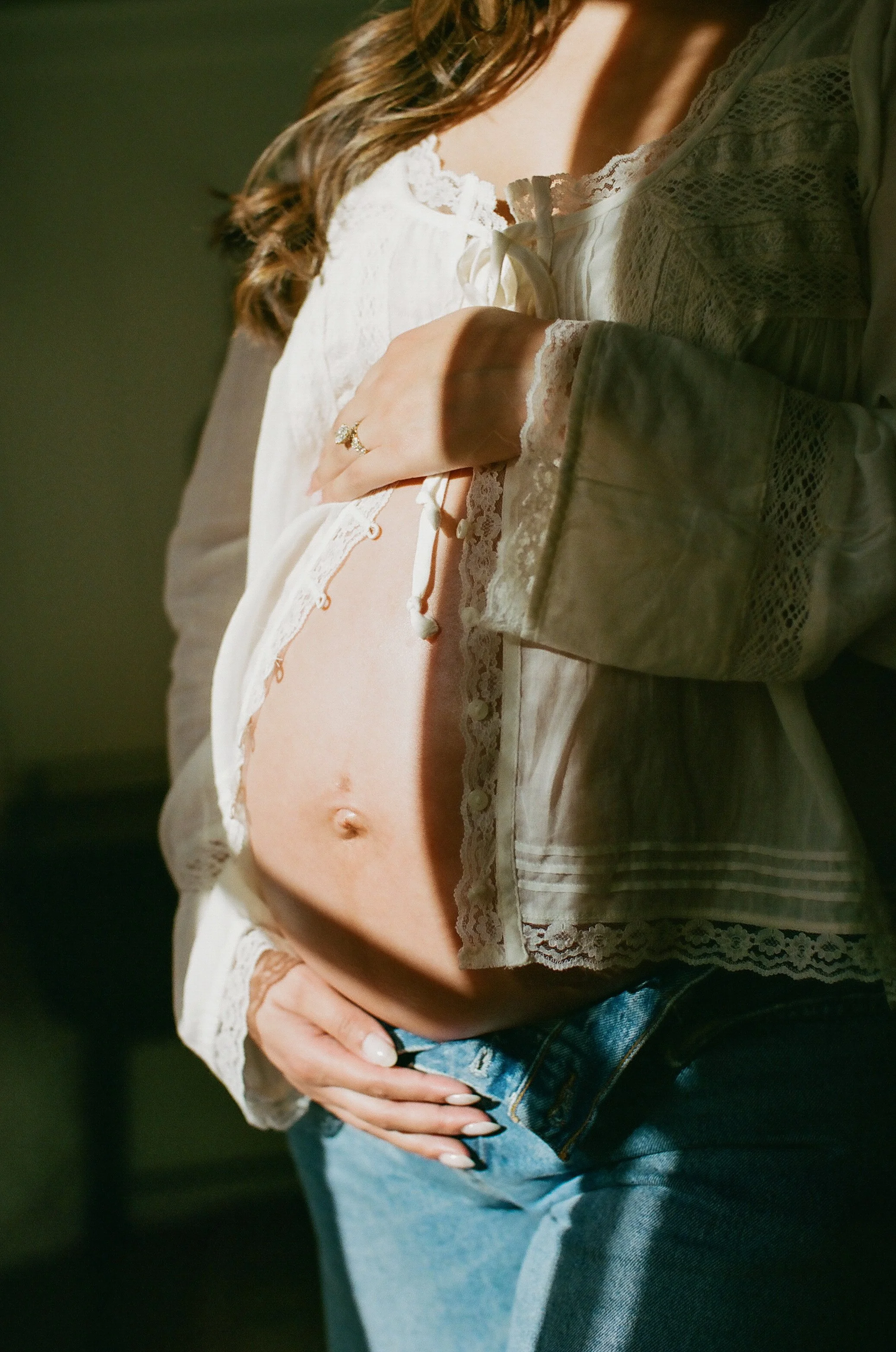 A woman wearing a white lace-trimmed blouse and blue jeans with one hand resting on her hip, showing a ring and her midriff.