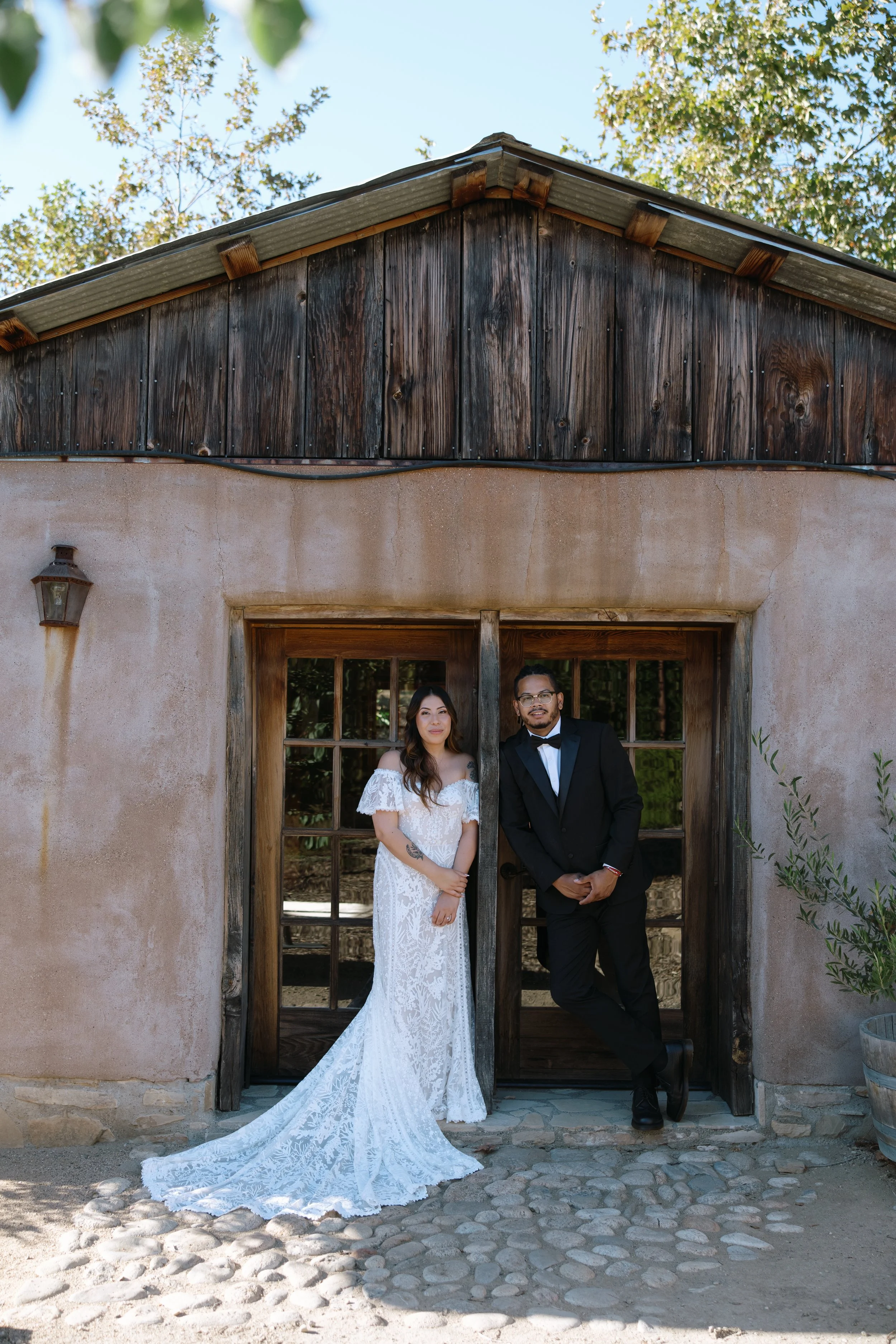 A man and a woman dressed in formal attire standing in front of a rustic wooden and stucco door with a window. The woman is wearing a white lace wedding dress, and the man is wearing a black tuxedo. They are standing close together outdoors, with tre