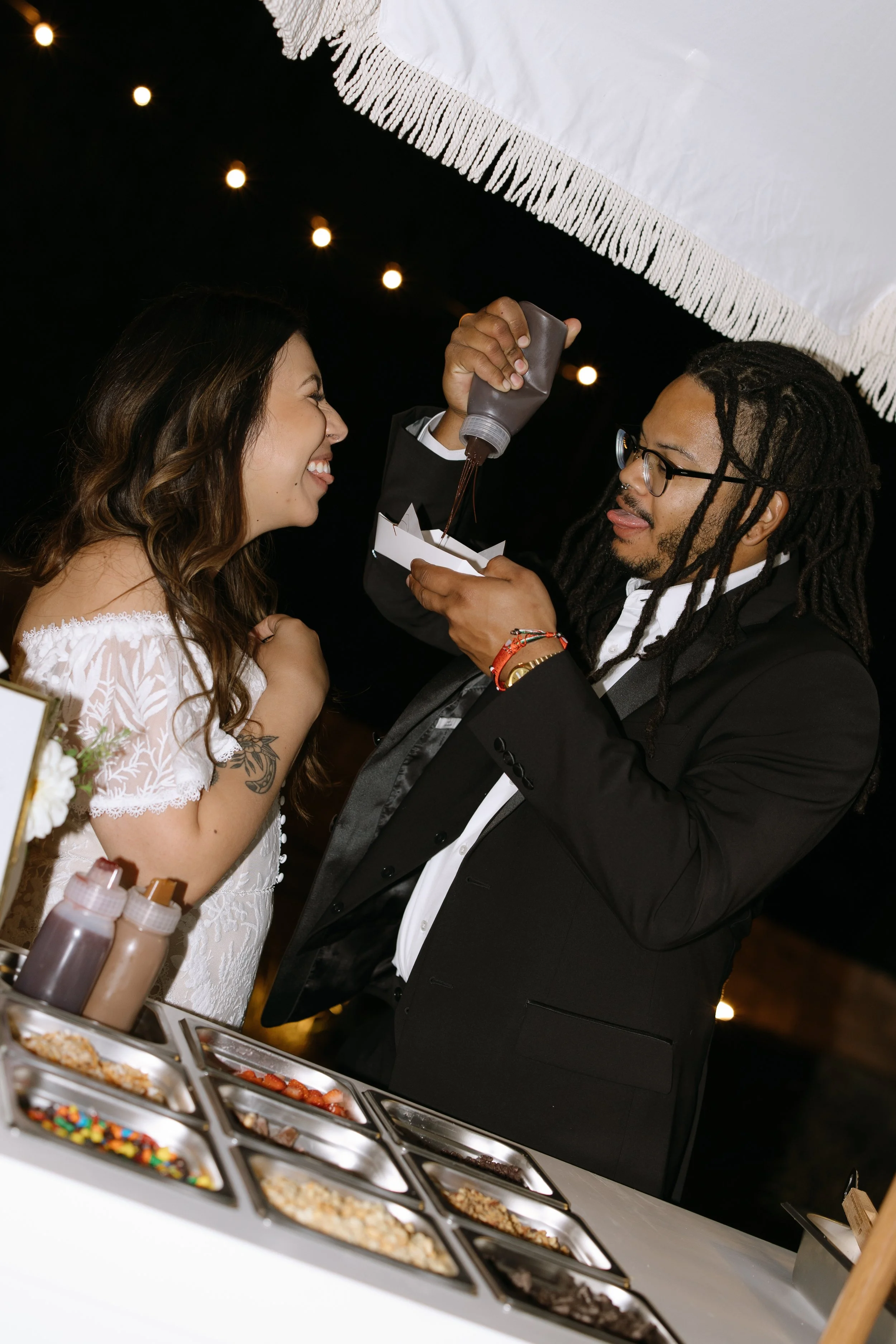 A bride and groom smiling and looking at each other during their wedding reception. The groom is putting chocolate syrup on a plate as part of a cake decorating activity. There is a table with various toppings such as candies, nuts, and sprinkles in 
