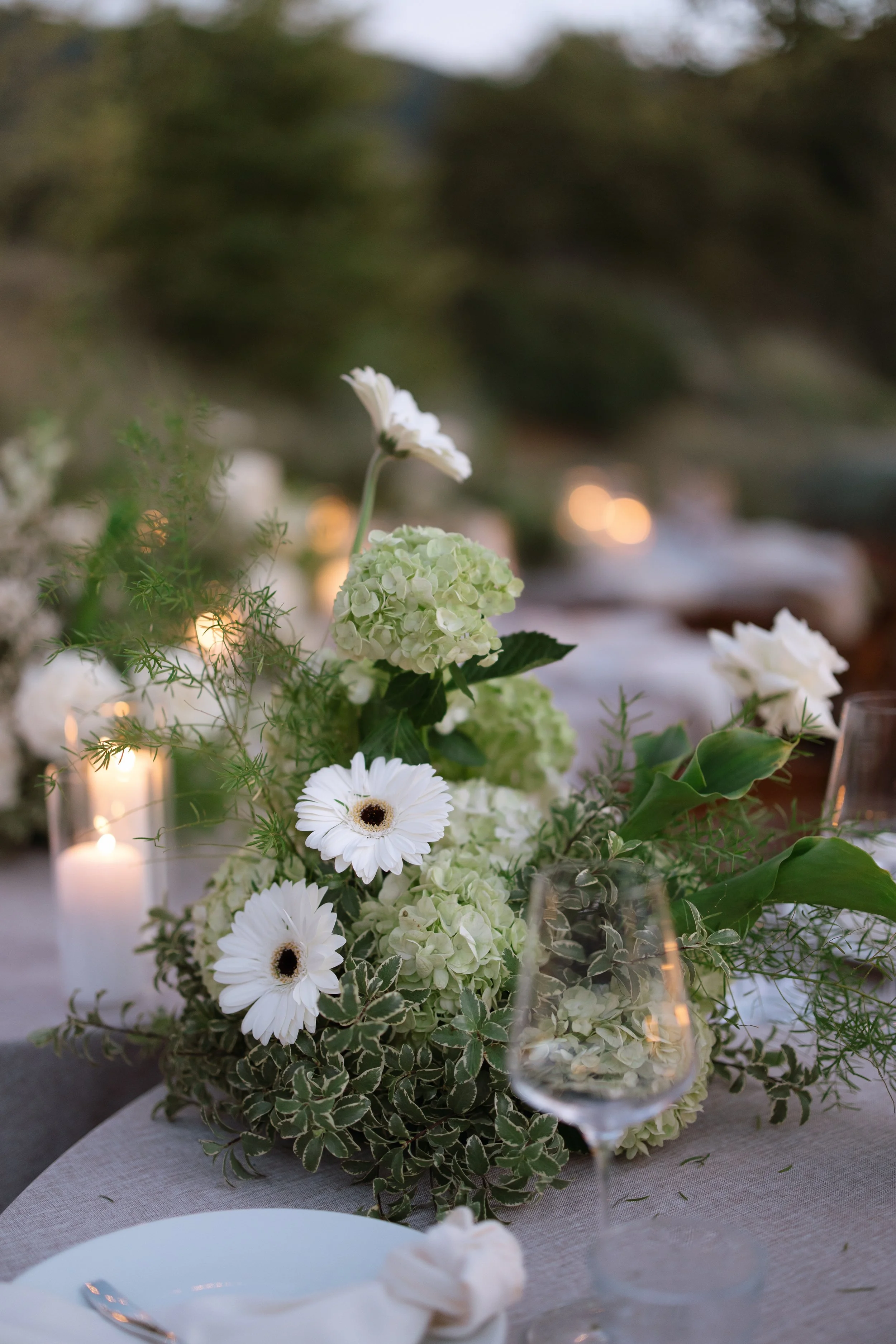 Elegant floral centerpiece with white daisies, green hydrangeas, and greenery on a table with glassware and candles, set in an outdoor or garden setting.