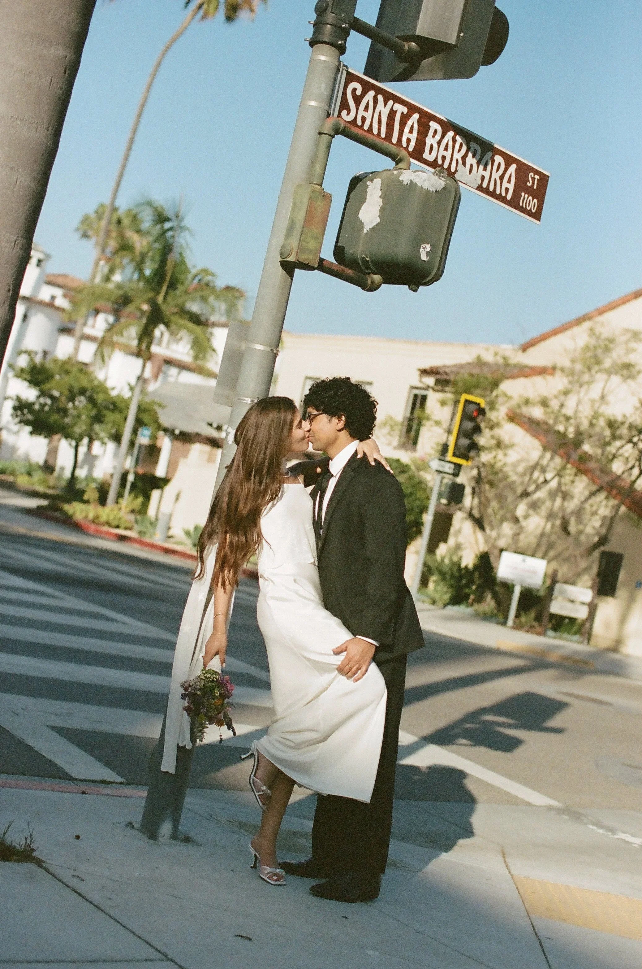 A newly married couple kissing at a crosswalk in front of a street sign that reads 'Santa Barbara St'. The bride is wearing a white dress and heels, holding a bouquet, and the groom is in a black suit and tie. They are standing close together, with the bride one leg slightly raised, showing their romantic moment. The background includes palm trees, buildings, and traffic lights.