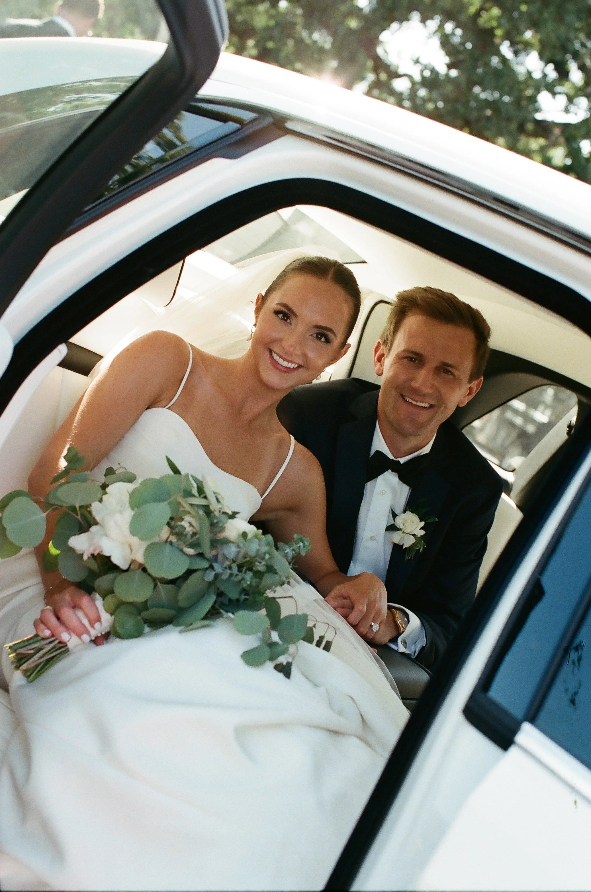 A smiling bride and groom sitting inside a car, dressed in wedding attire, with the bride holding a bouquet of flowers.