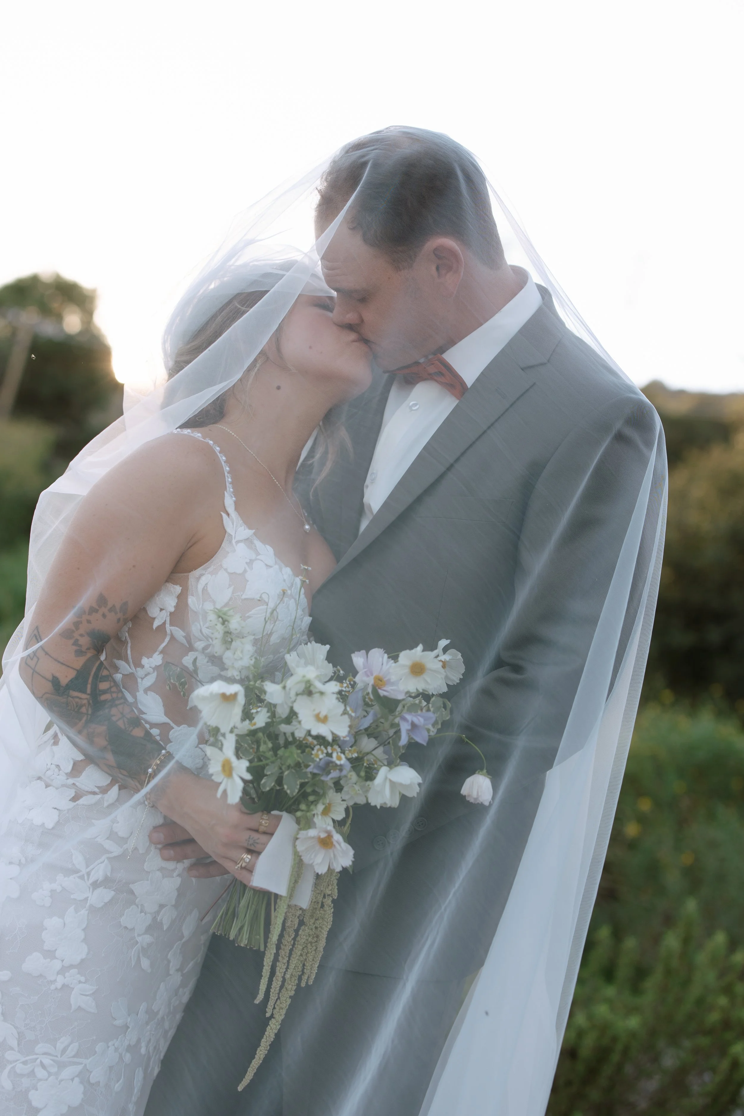 A bride and groom kissing under a wedding veil outdoors, with the bride holding a bouquet of flowers and smiling.