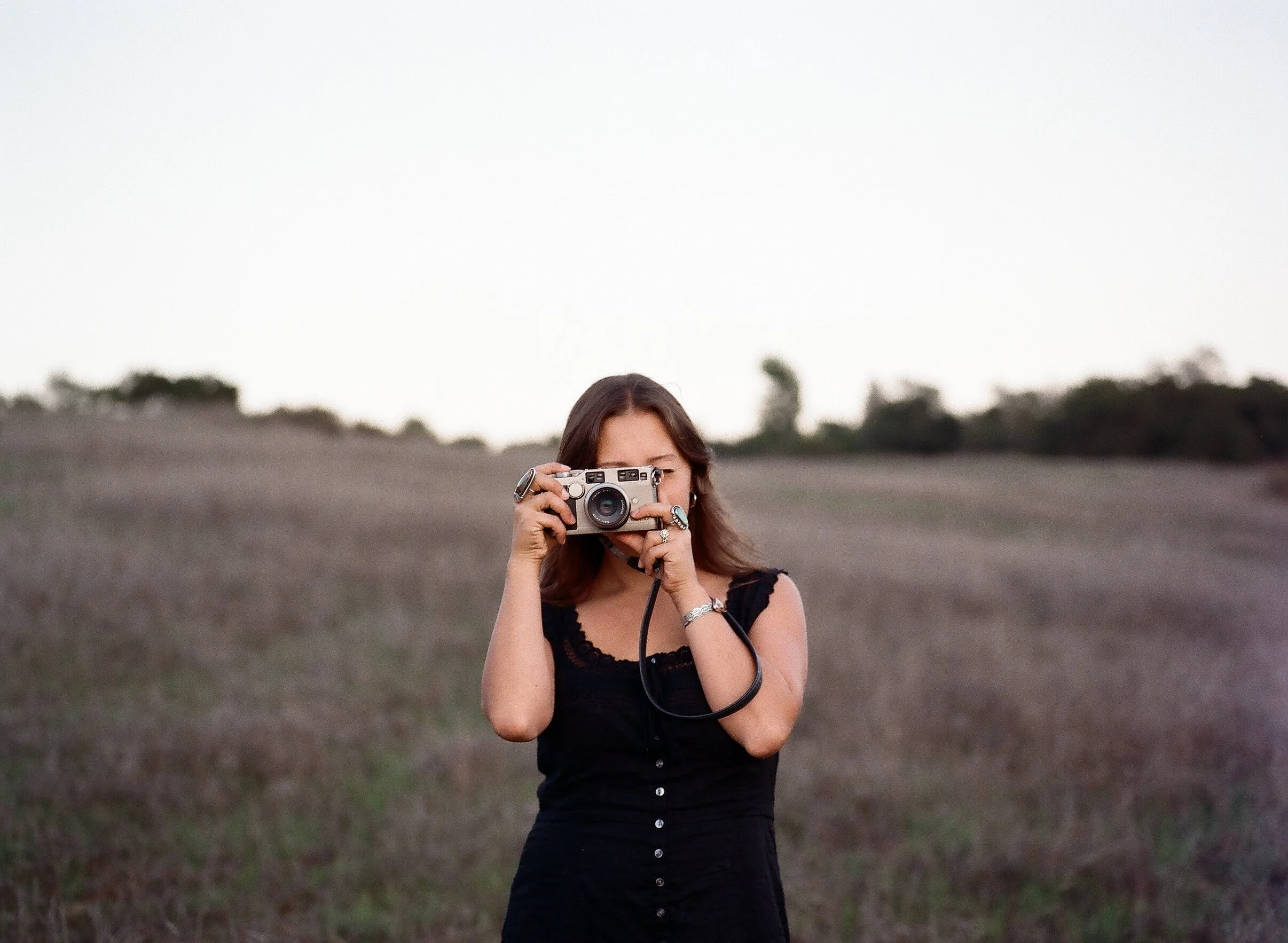 A woman with brown hair, wearing a black top, holding a vintage camera in front of her face, standing outdoors in a field with trees in the background during dusk.