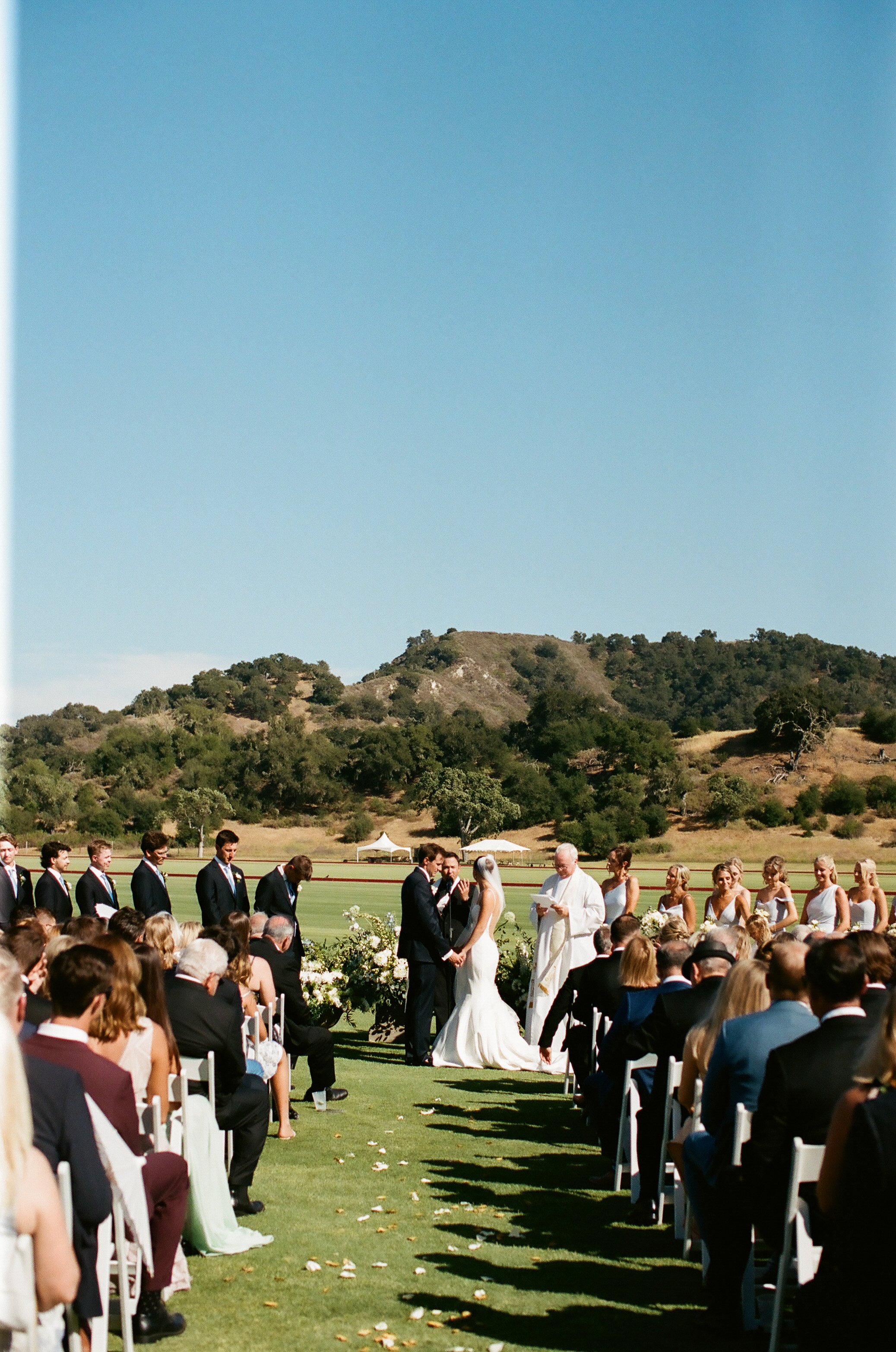 A wedding ceremony taking place on a sunny day outdoors. The bride and groom stand before an officiant, with the audience seated on either side. The background features rolling hills and a clear blue sky.