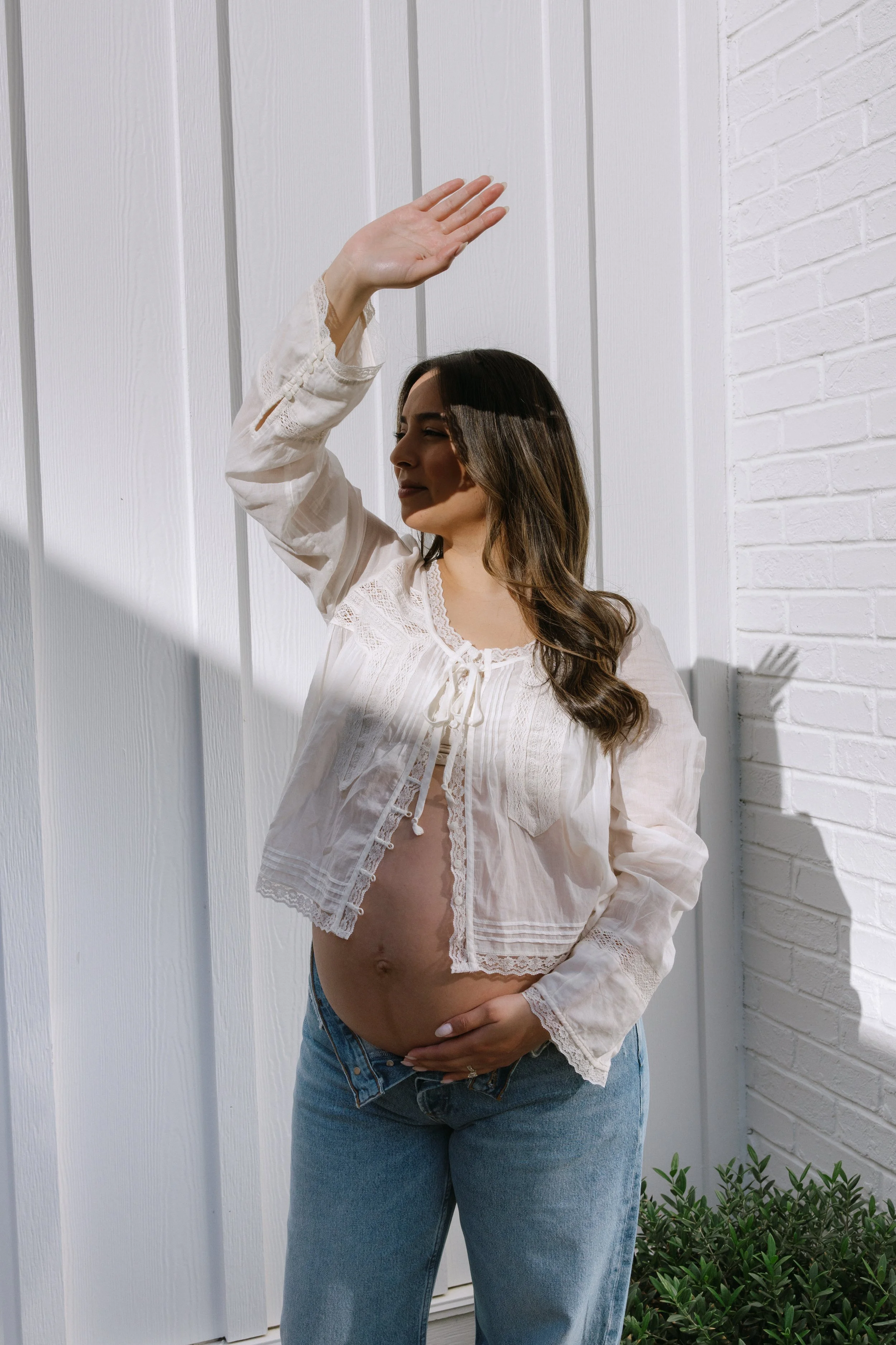 Pregnant woman wearing a white lace blouse and blue jeans, standing outdoors against a white paneled and brick wall, cradling her belly with one hand and raising the other hand in front of her face.