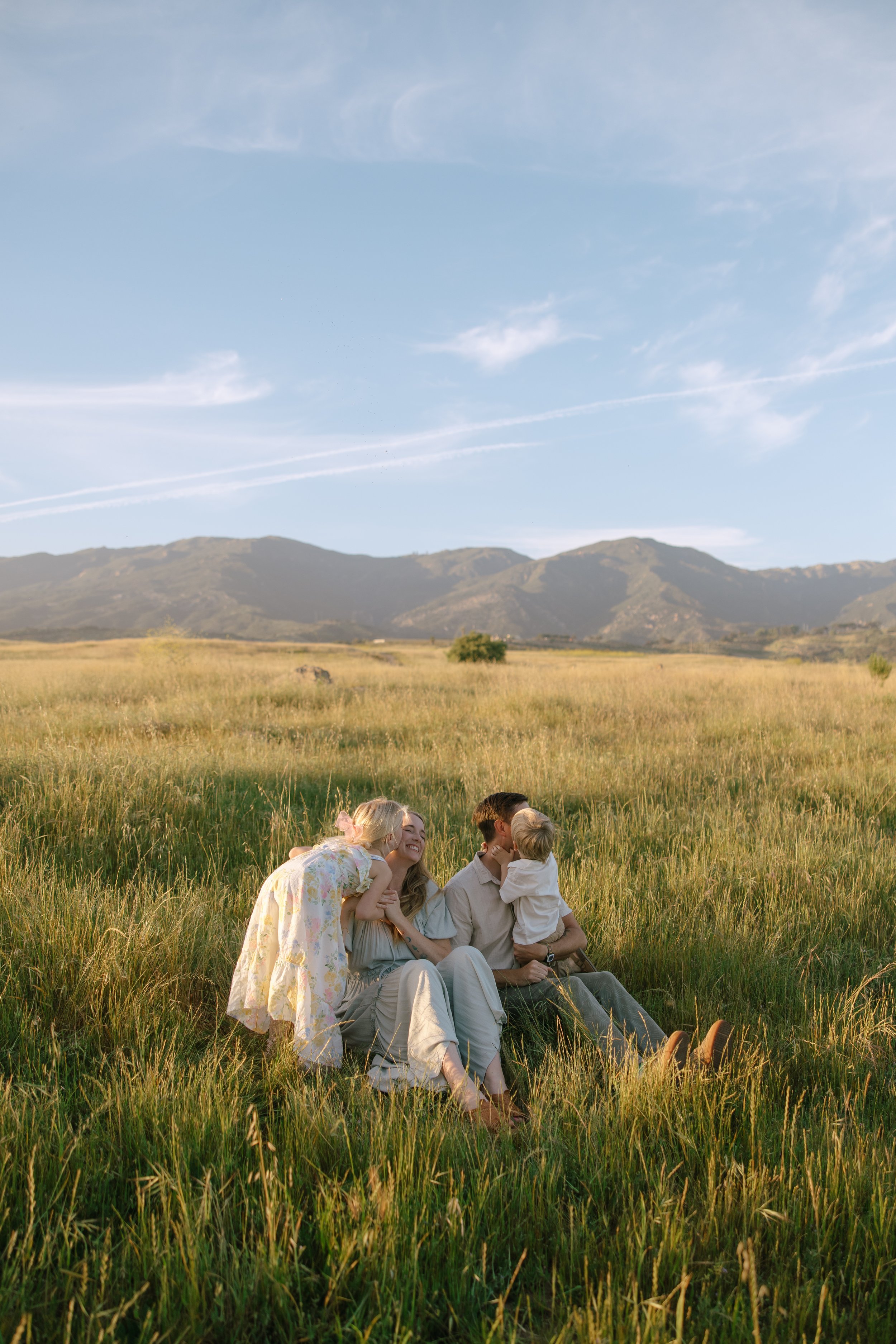 A family of four, including a woman, a man, a young girl, and a young boy, sitting and playing in a grassy field with mountains in the background on a clear sunny day.