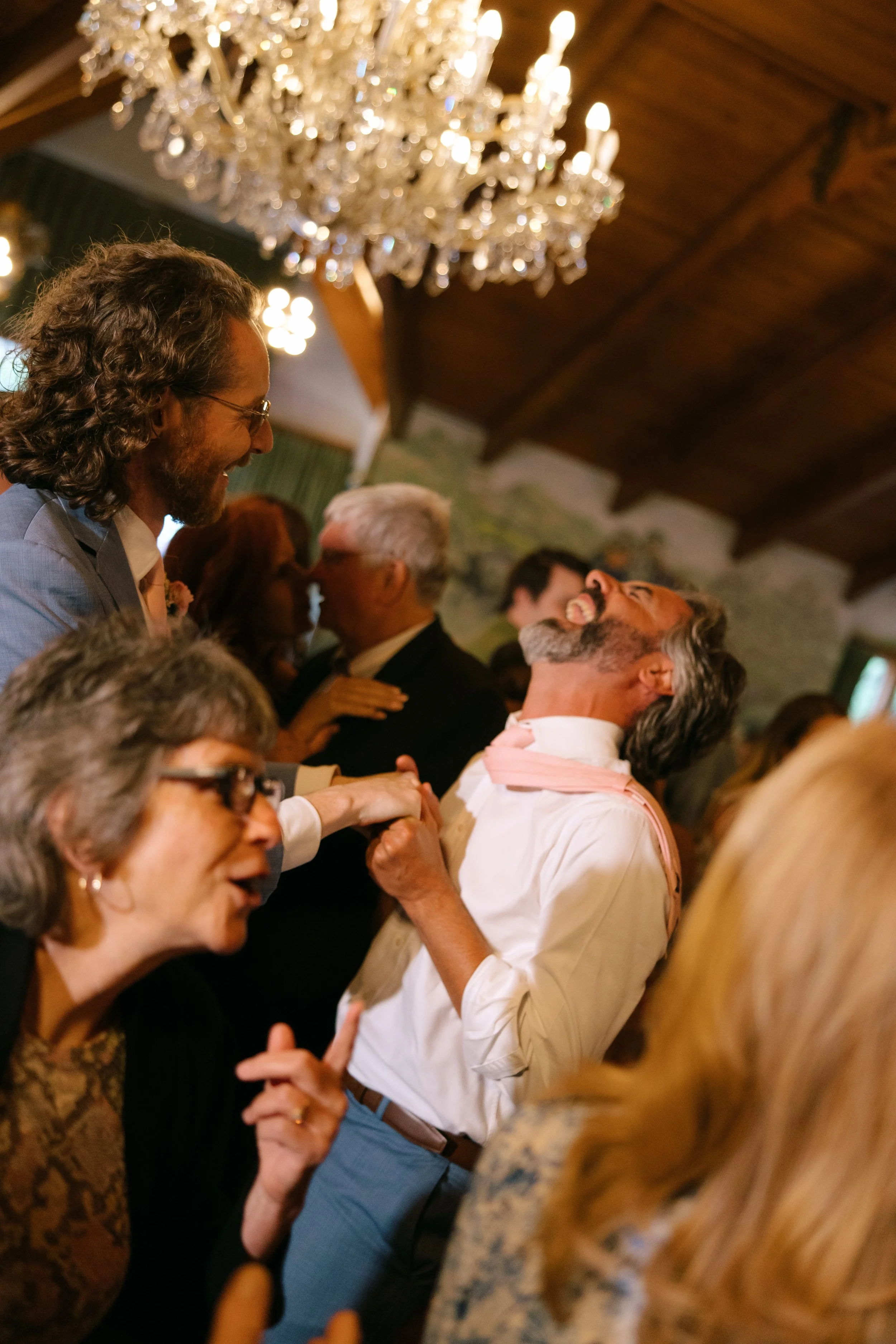 People dancing and enjoying a celebration in a room with a chandelier and a wooden ceiling.