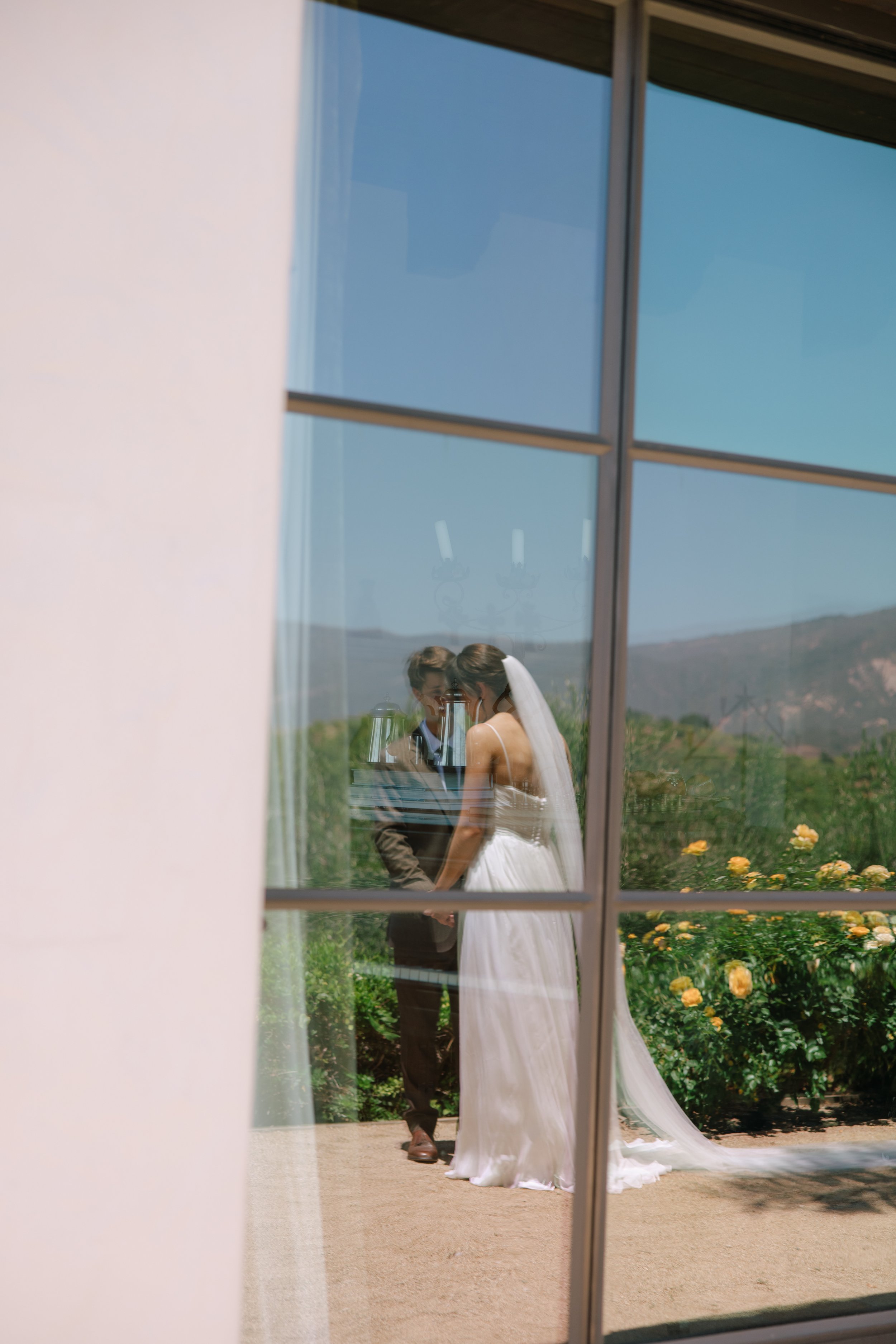 A bride and groom holding hands and kissing outside, viewed through a window with a white frame, with a garden and mountain landscape in the background.