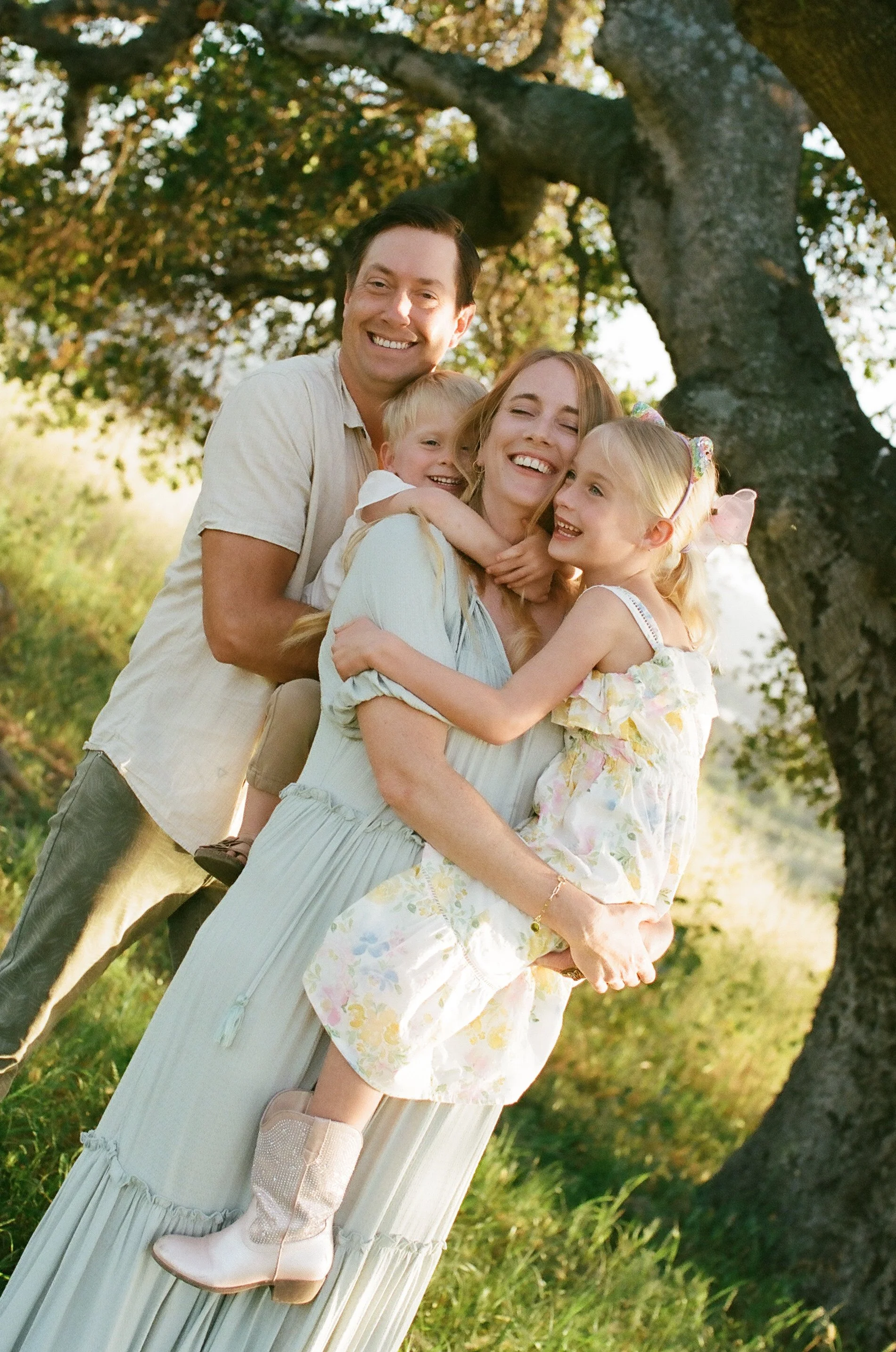 Family of four embracing outdoors under a large tree during daytime, smiling and showing affection.