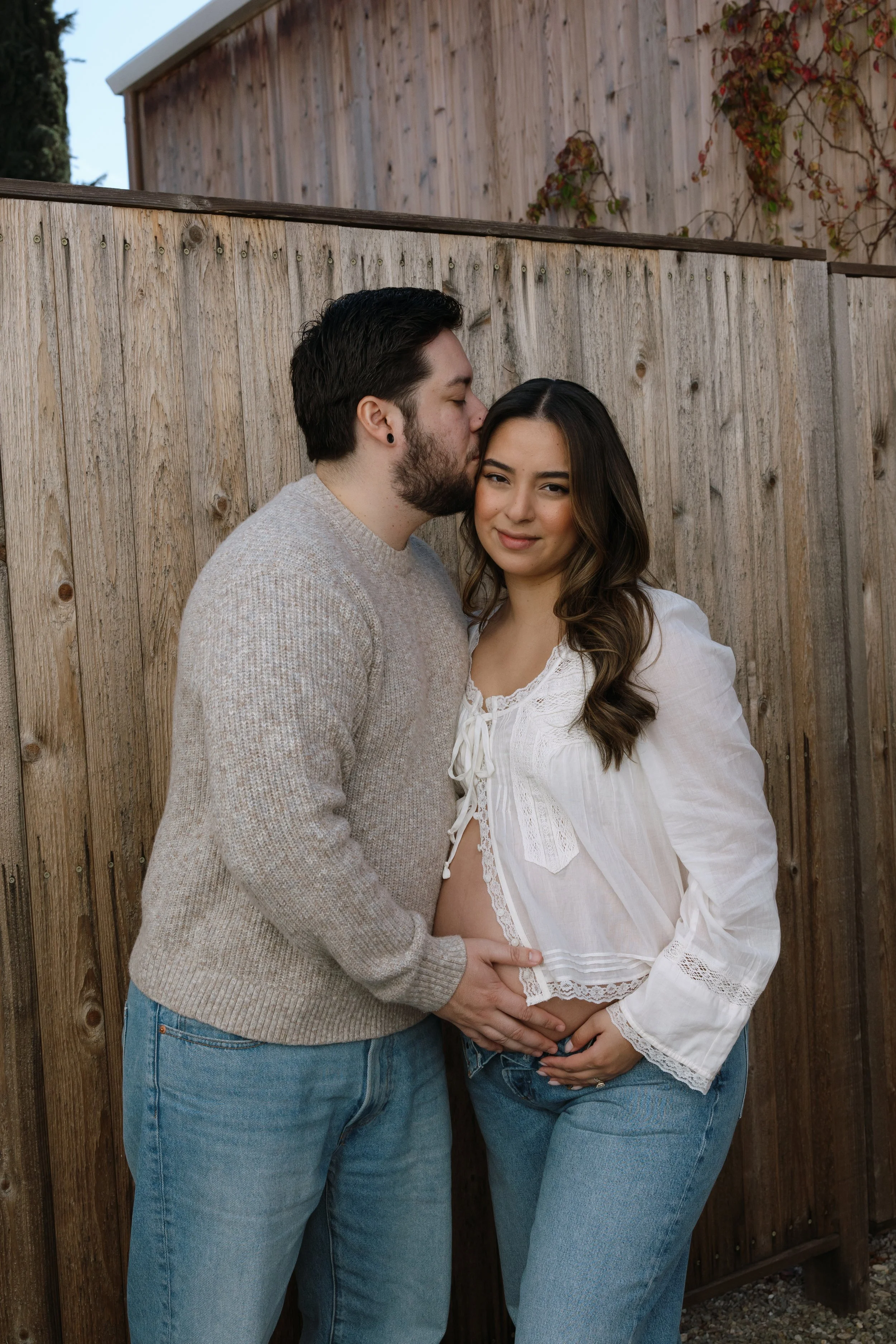 A pregnant woman and a man kissing on the side of a wooden fence outdoors. The woman is wearing a white blouse and jeans, and the man is in a gray sweater and jeans. They are holding her belly and standing close together.