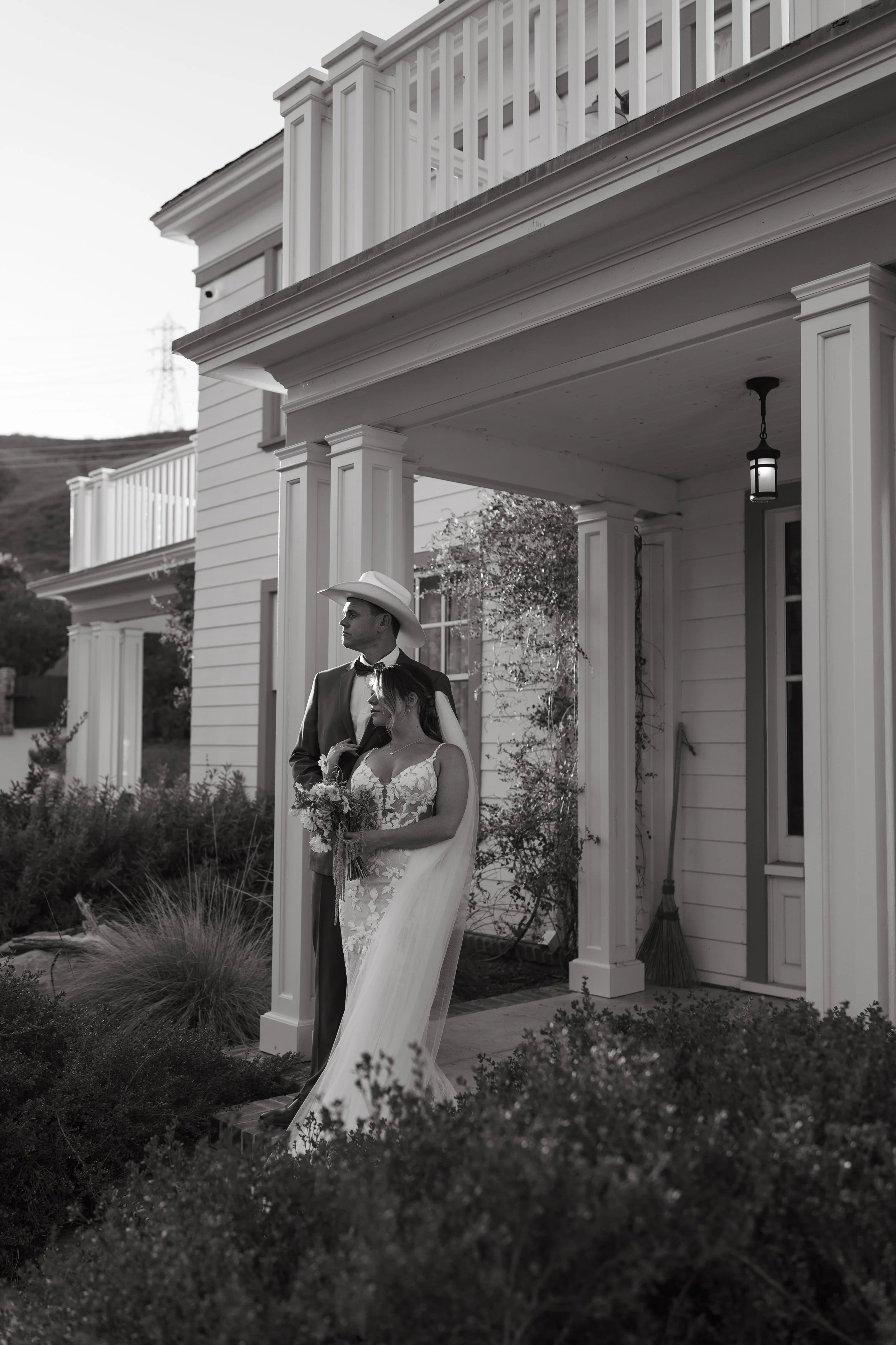 A black and white photo of a newlywed couple standing outside a white house with large columns, a porch light, and a balcony. The groom is wearing a suit and cowboy hat, and the bride is in a lace wedding gown holding a bouquet.