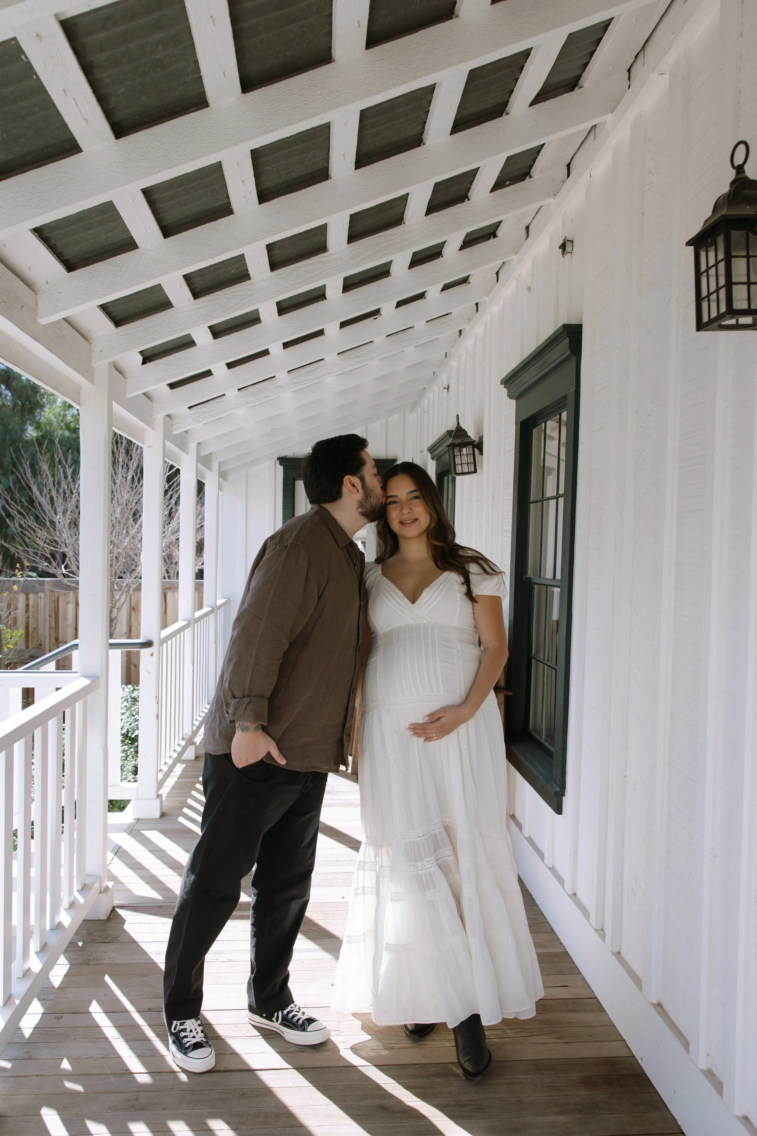 A man kisses a woman on the forehead on a porch, with the woman holding her pregnant belly. The porch has white wooden beams and walls, black window frames, and lantern-style lights.